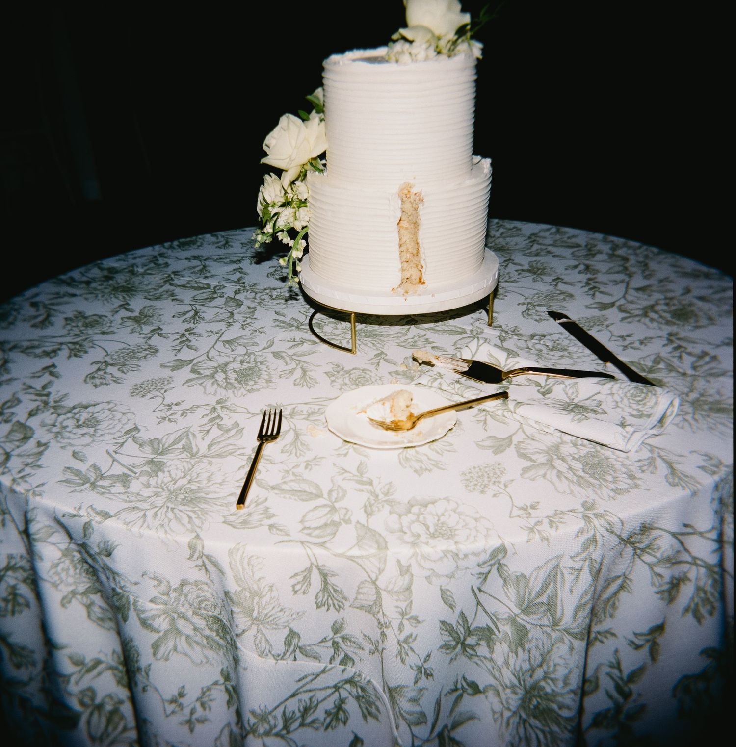 Simple white tiered wedding cake on a patterned tablecloth with scattered utensils.