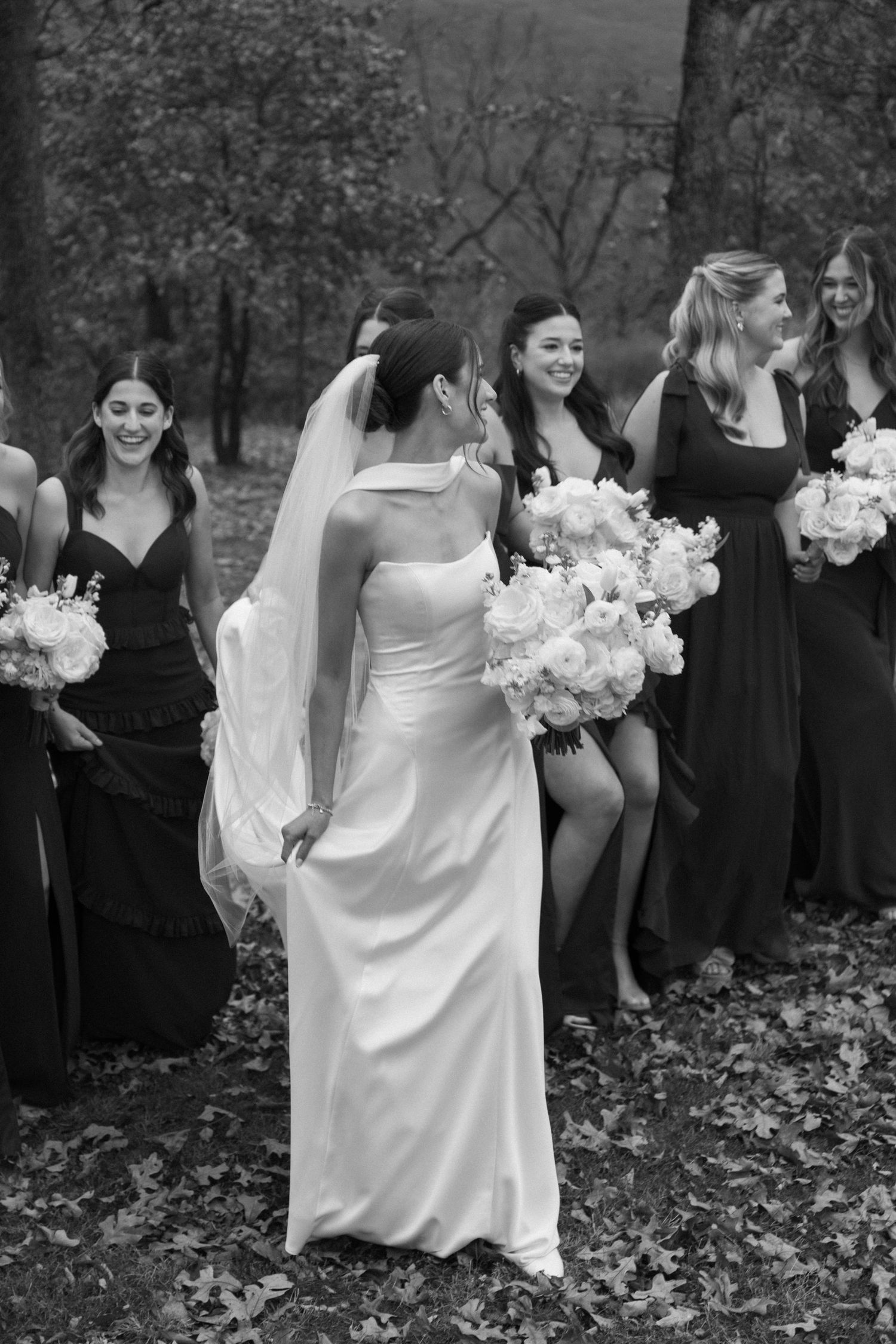 Wedding party in black and white attire poses outdoors among fallen autumn leaves.