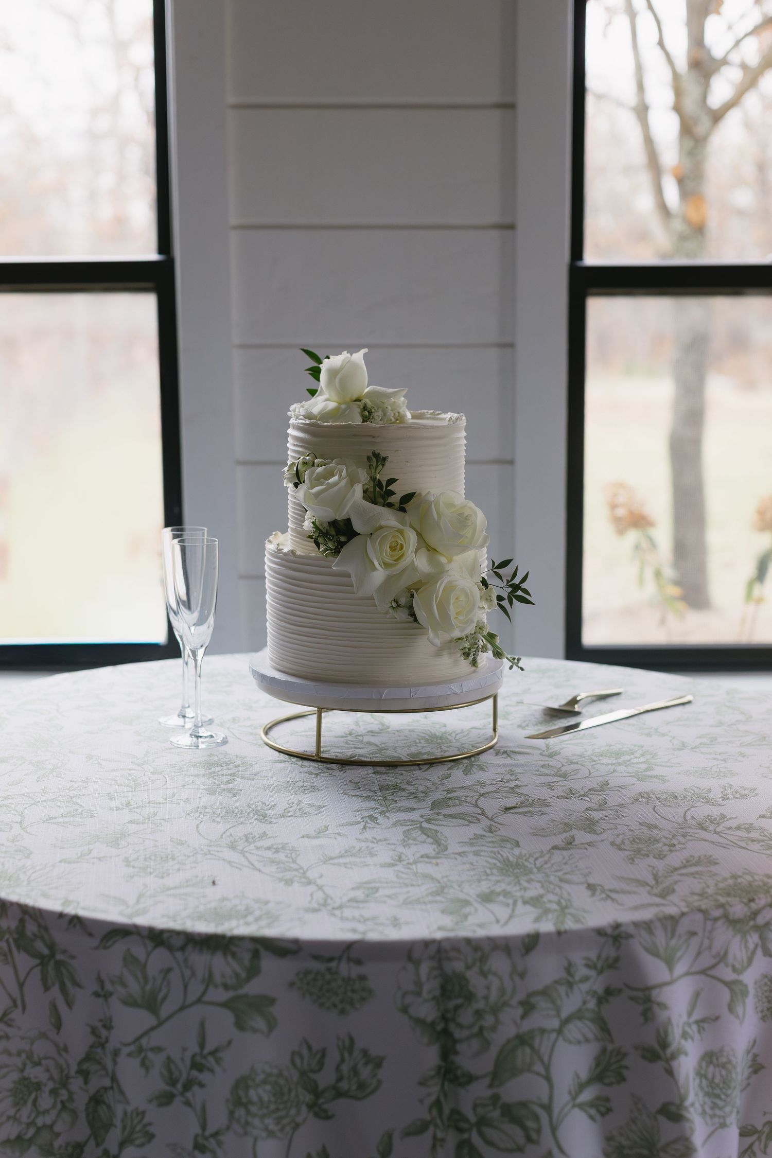 Elegant white wedding cake with fresh roses displayed on a floral patterned tablecloth near windows.