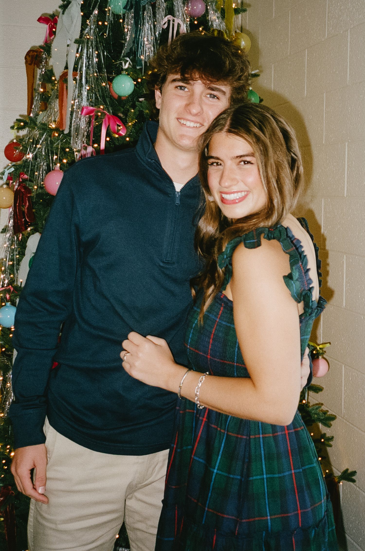 A couple poses together in front of a decorated Christmas tree, wearing coordinating navy and plaid holiday outfits.