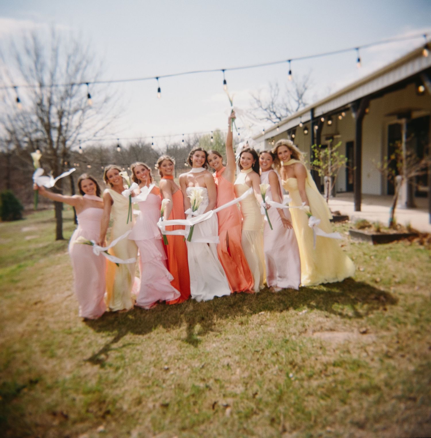 Wedding party in coral and pink dresses poses together on a grassy hillside with string lights overhead.