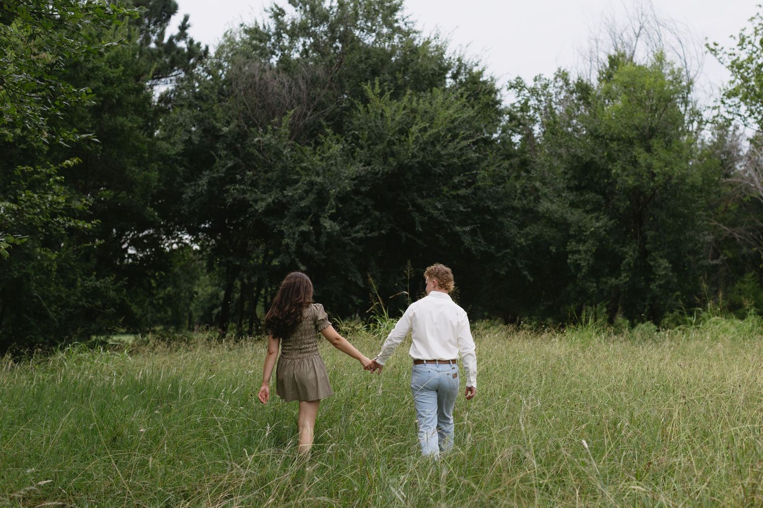 Two figures walk through a distant meadow surrounded by green trees.