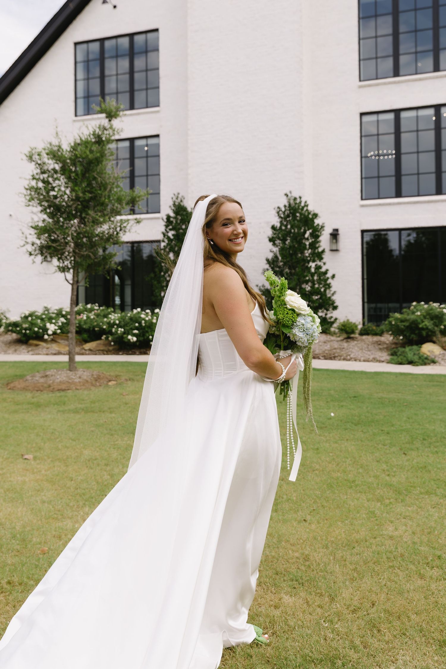 Bride in classic white gown poses outdoors on lawn near modern building with large windows.