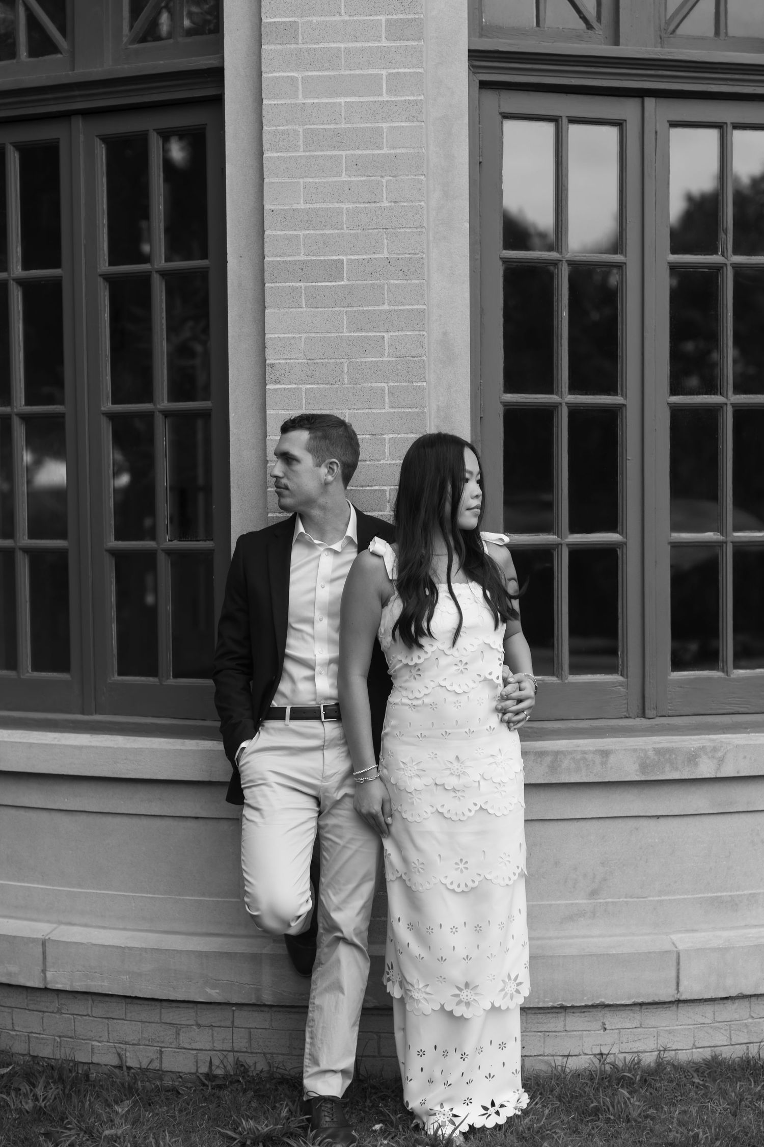 A stylish couple poses against a gray brick building with large windows during their engagement session.