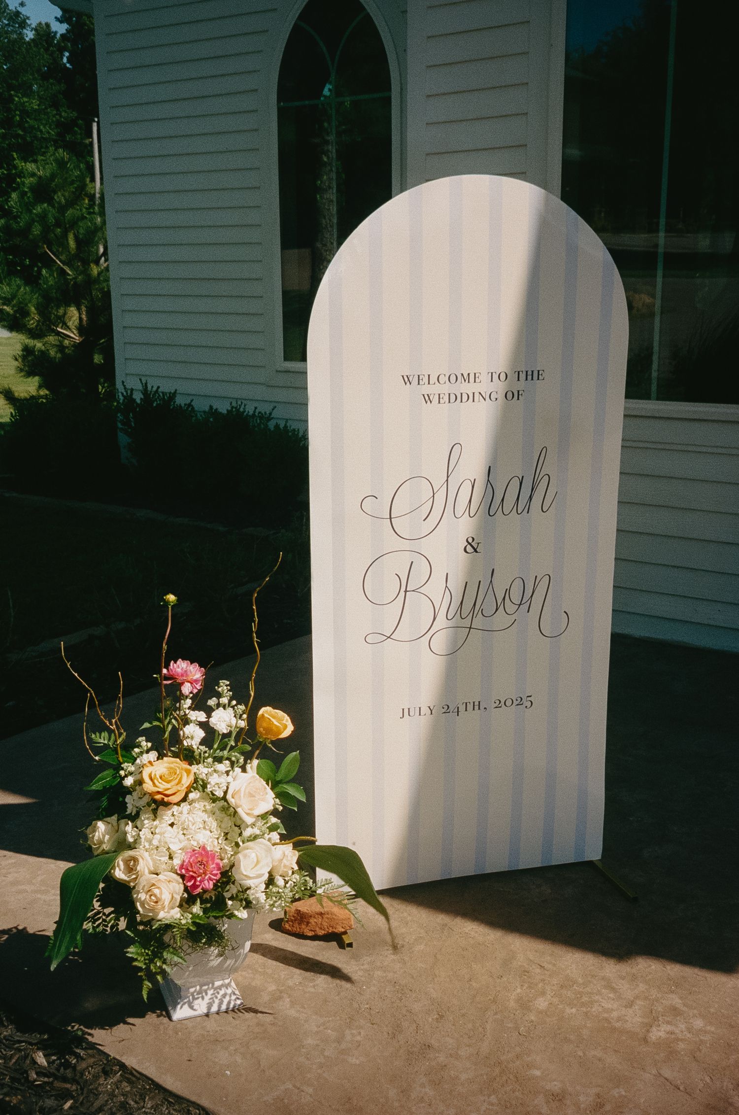 Elegant wedding welcome sign with floral arrangement featuring pink, yellow and white flowers outside a white church building.