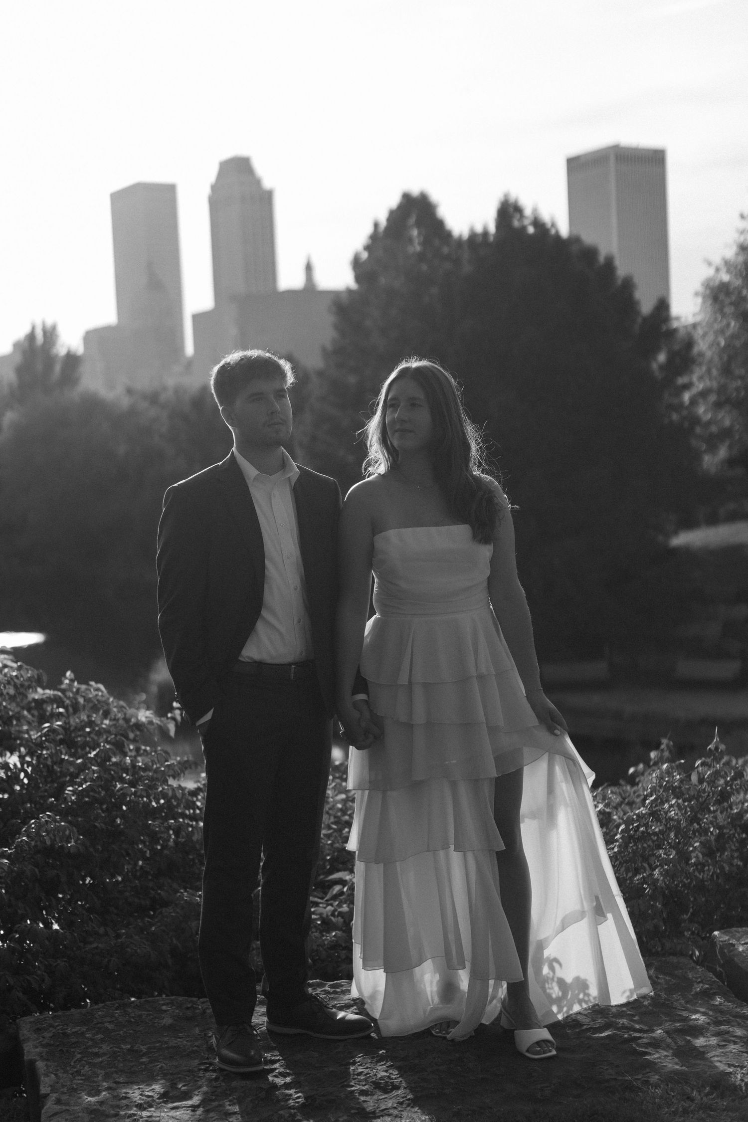 A silhouetted couple poses against a city skyline at sunset in formal attire.