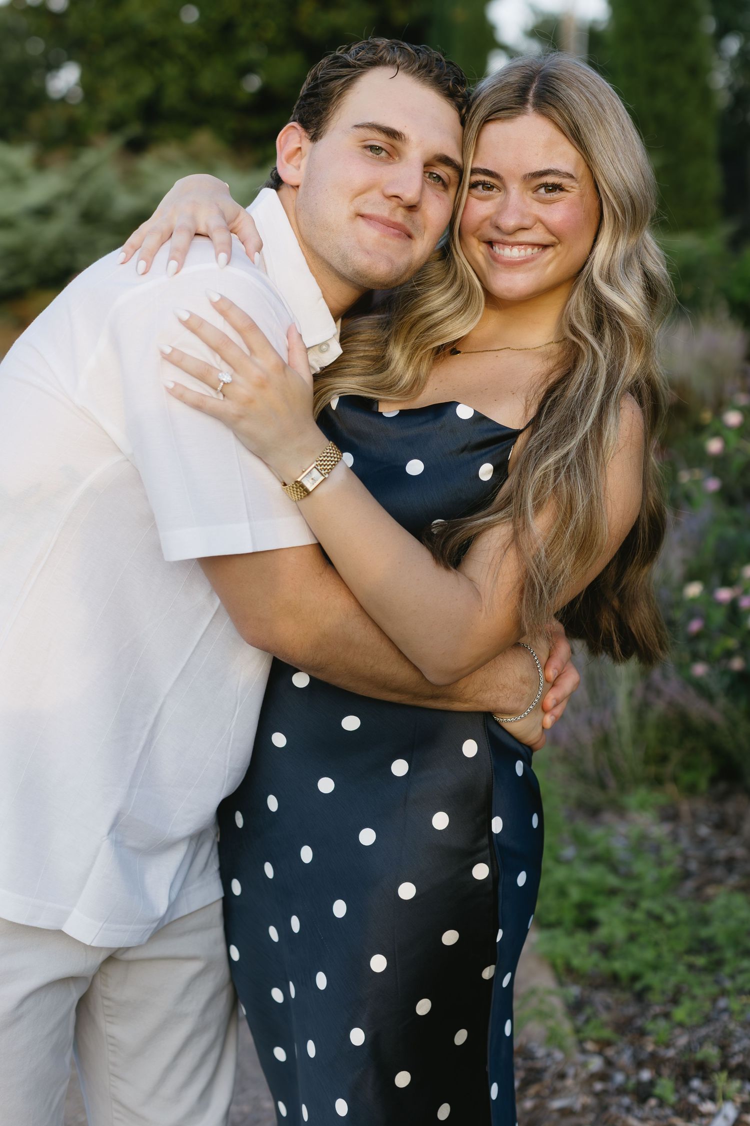 A happy couple embraces outdoors, wearing white and polka dot outfits against a garden background.