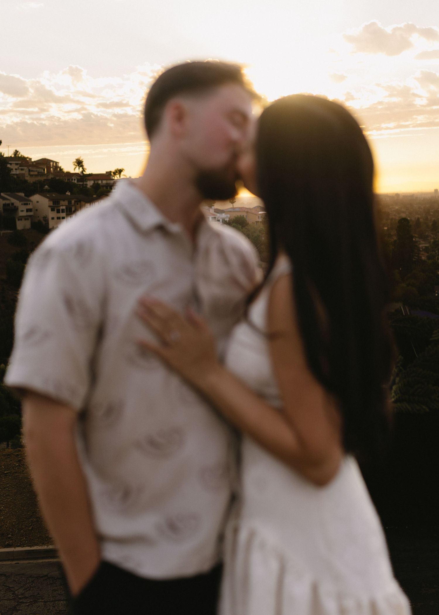 A romantic couple shares a tender kiss against a scenic sunset backdrop on a hilltop.