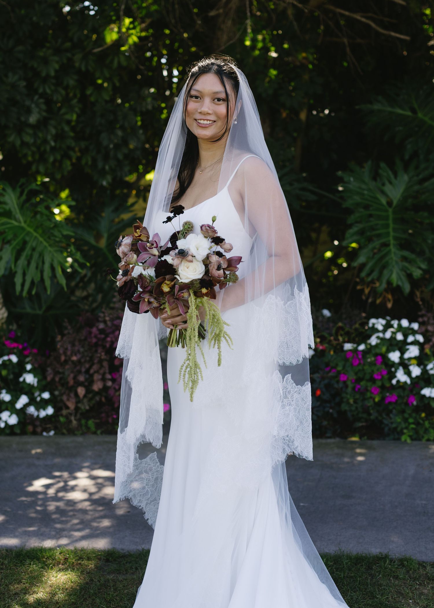 A bride in a white wedding gown holds a beautiful burgundy and white floral bouquet in an outdoor garden setting.