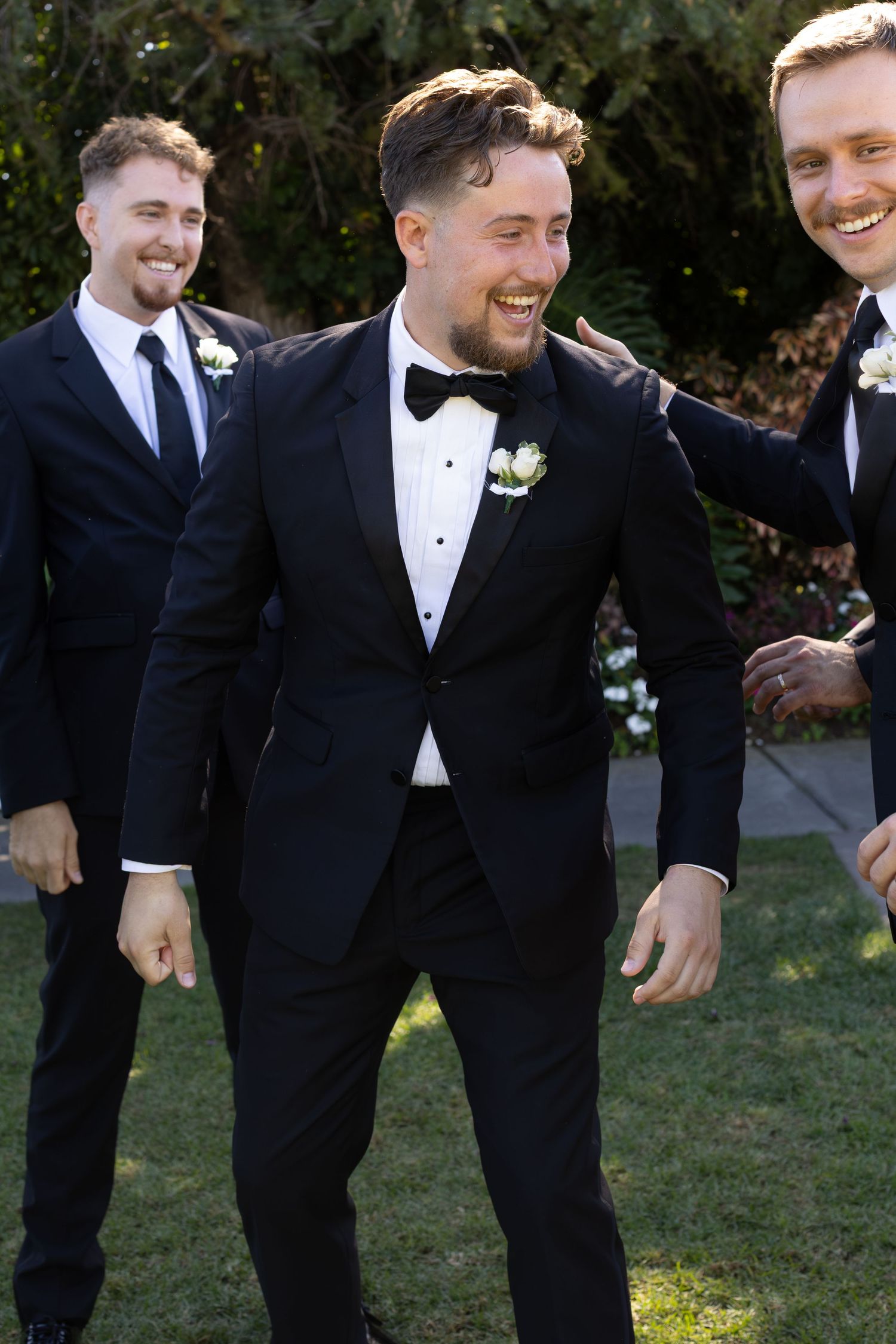 Groomsmen in black tuxedos share laughs and candid moments during outdoor wedding photography.