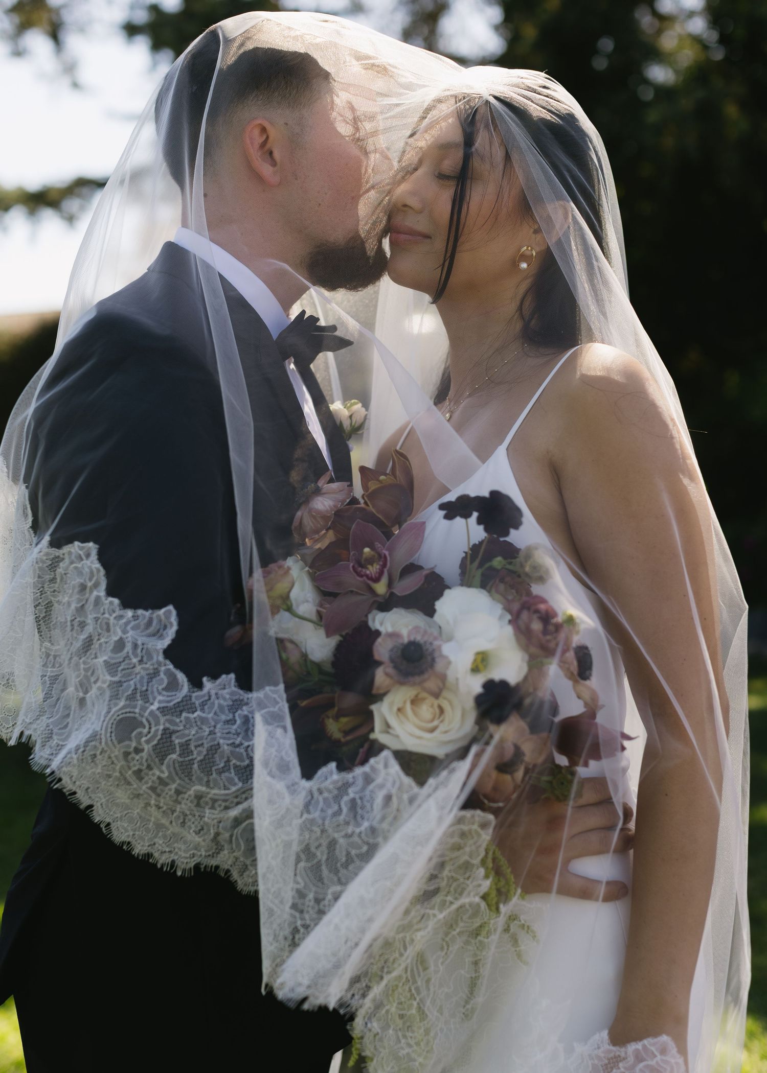 Romantic wedding moment with a couple embracing under a delicate lace veil while holding a purple and white bouquet.