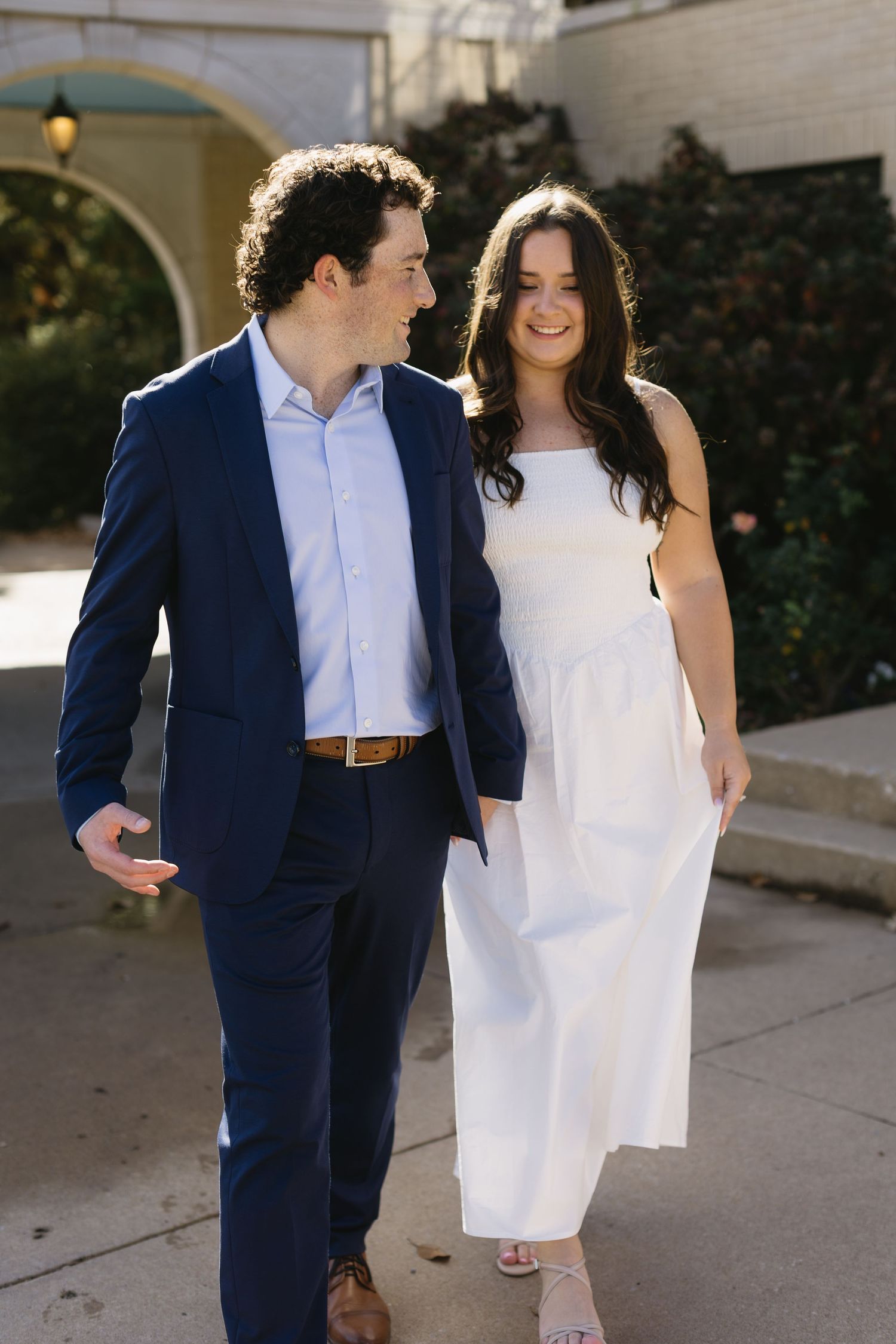 A couple in formal attire smiling while walking together, with her in white and him in navy blue.