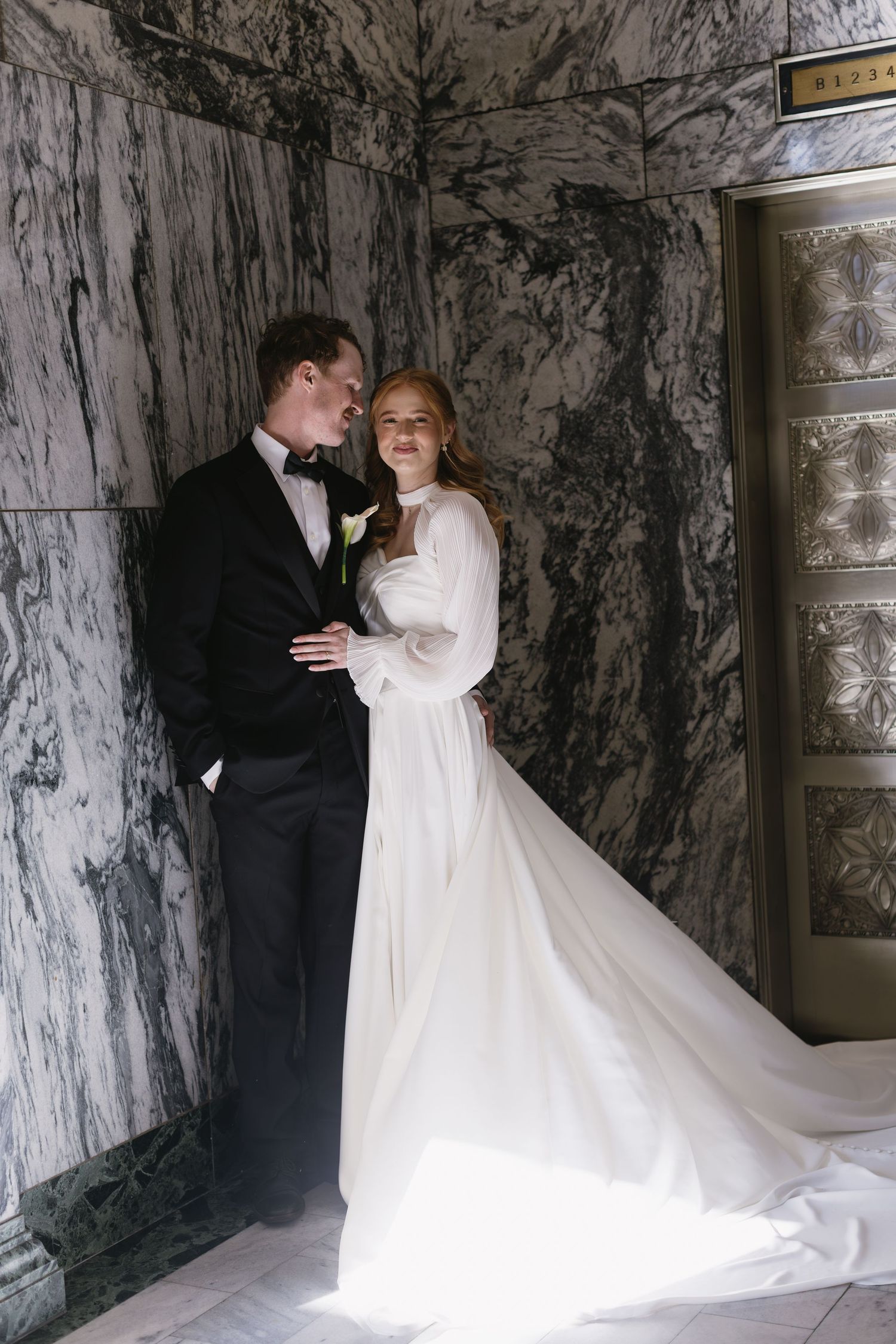 Wedding couple in elegant attire posing against marble walls in natural light.