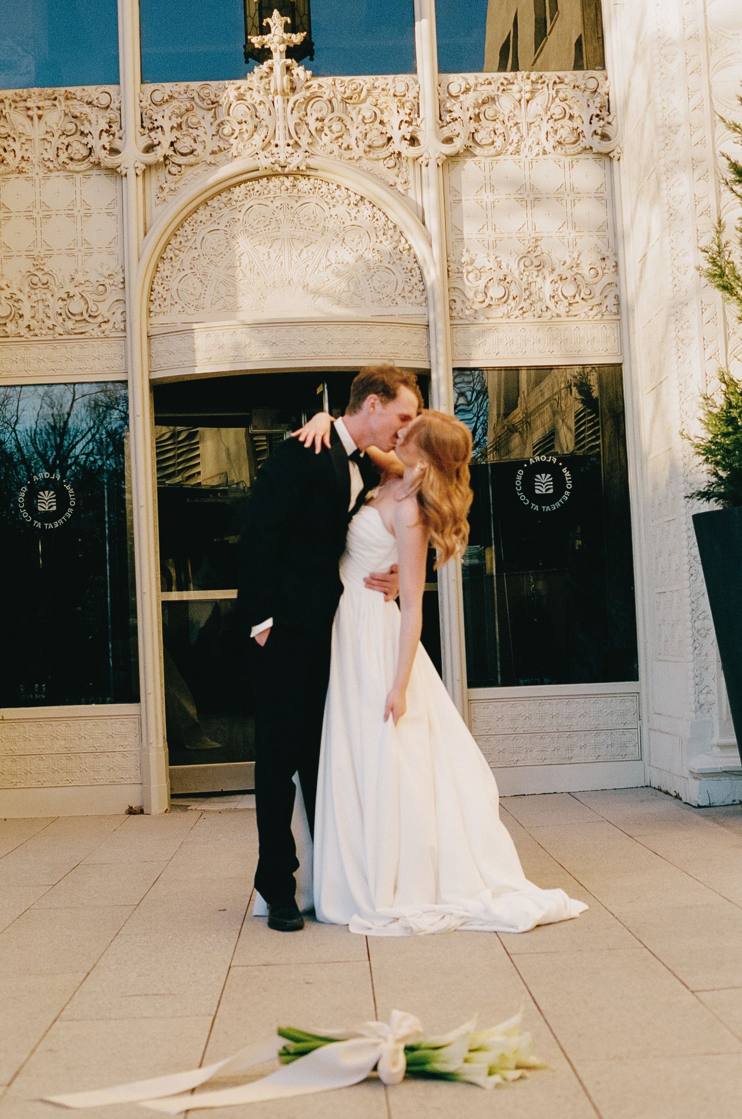 A romantic wedding portrait under an ornate white architectural arch with calla lilies on the ground.