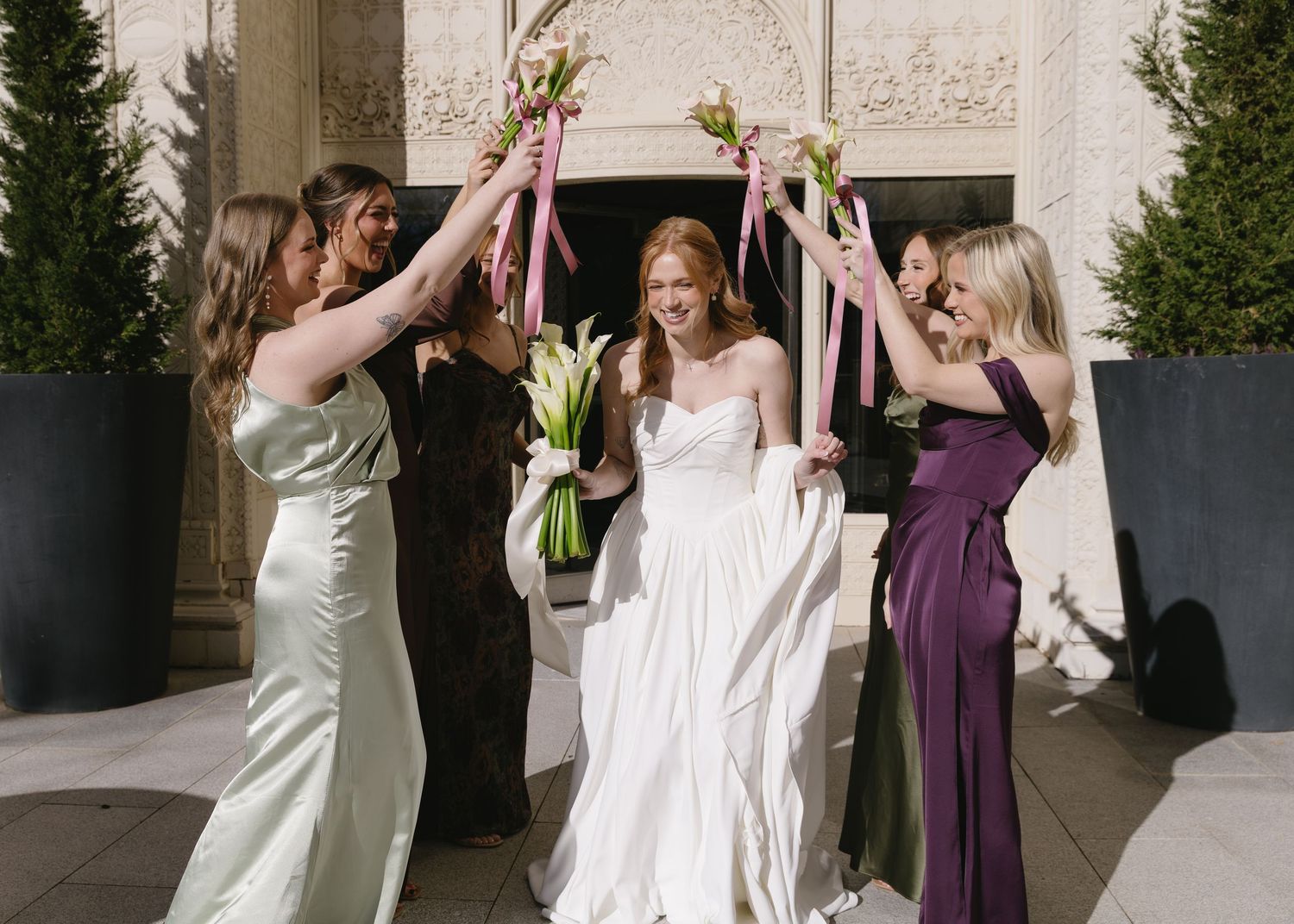 Wedding party celebrates with joyful poses and bouquets in an elegant outdoor setting.