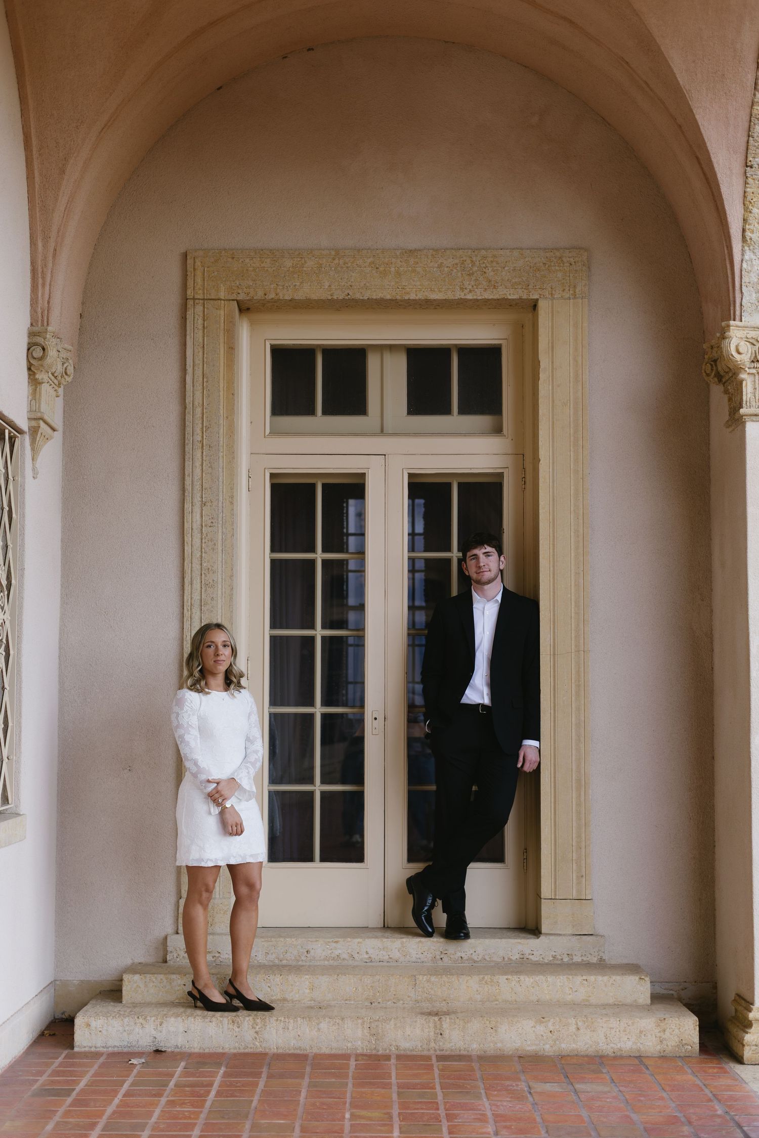 Couple in formal attire walking through an elegant arched hallway with terracotta floors and white columns.