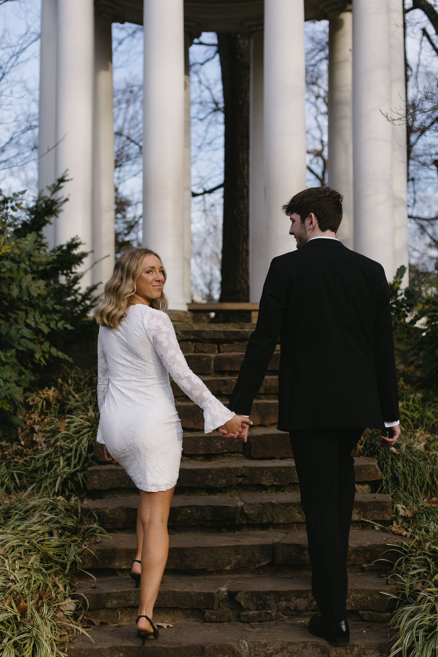 A couple walks up stone steps surrounded by white columns in formal attire.