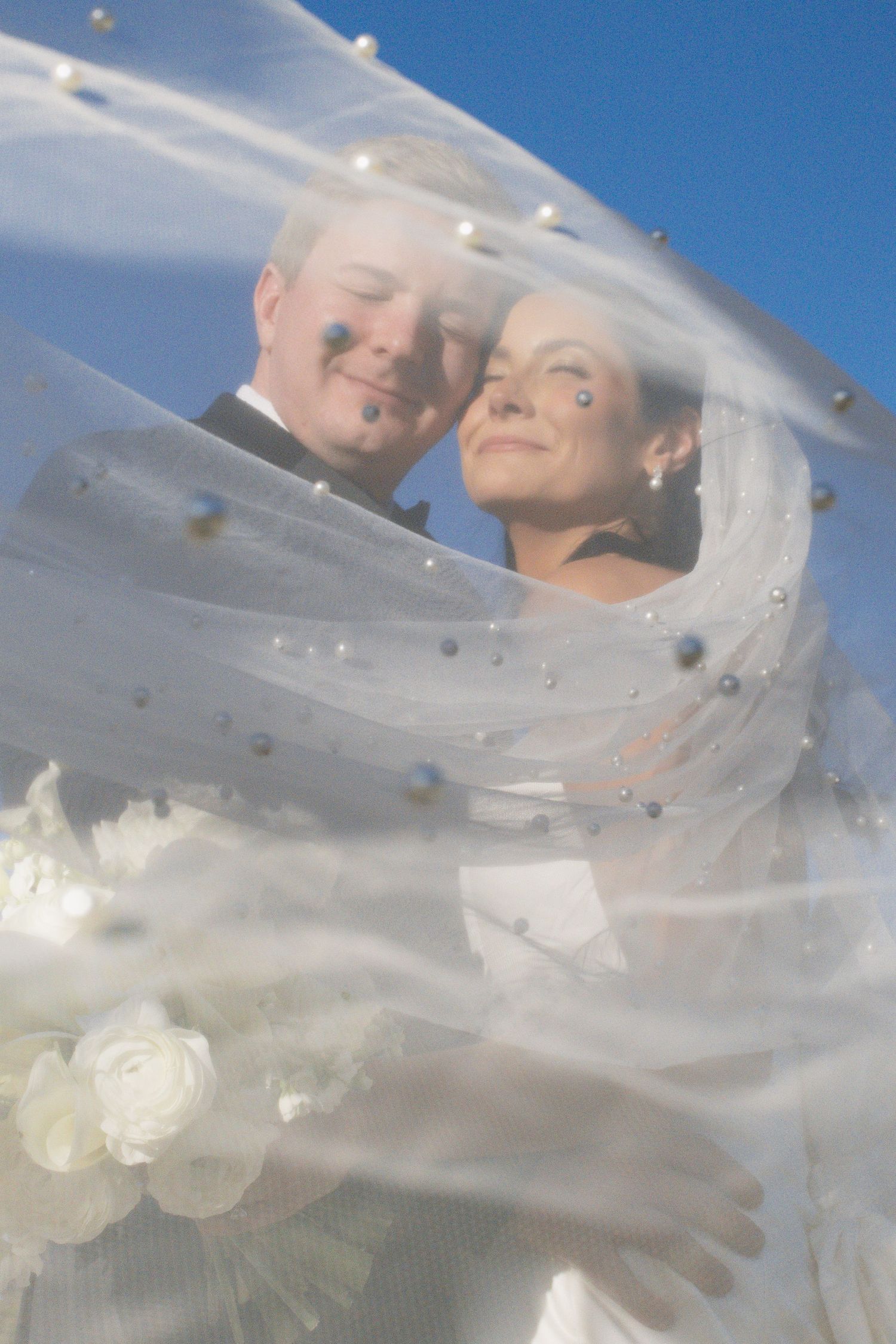 A wedding couple smiles through flowing white veil against blue sky.