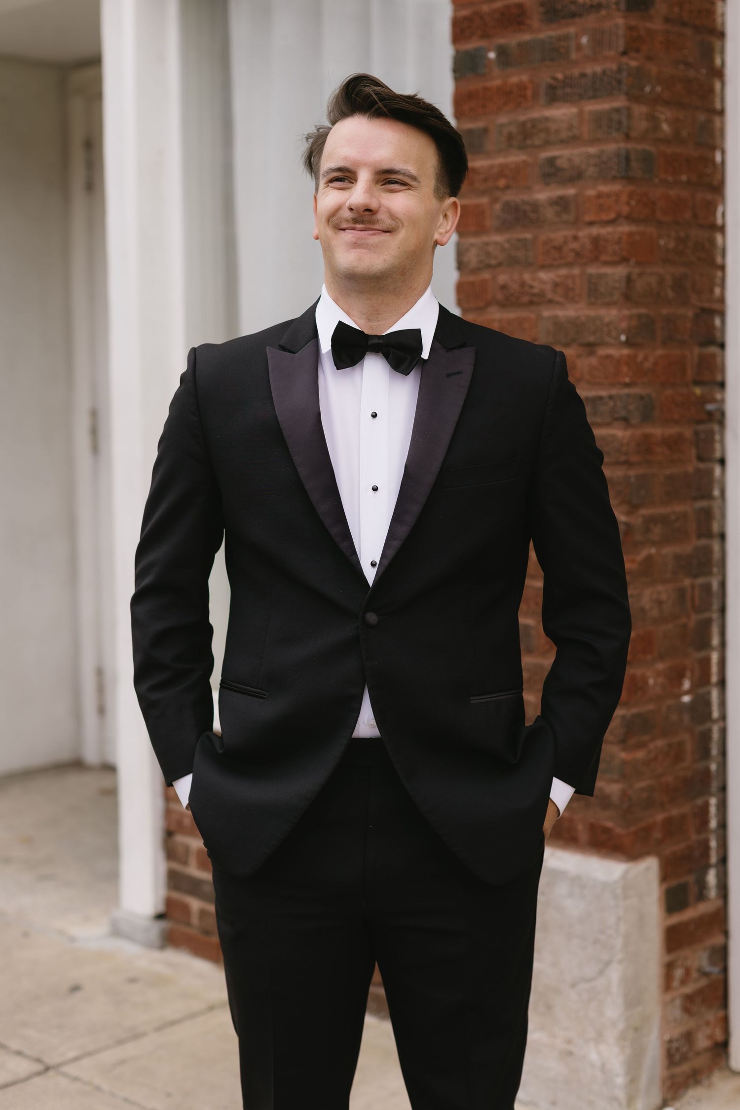 Groom in classic black tuxedo poses against brick wall columns.