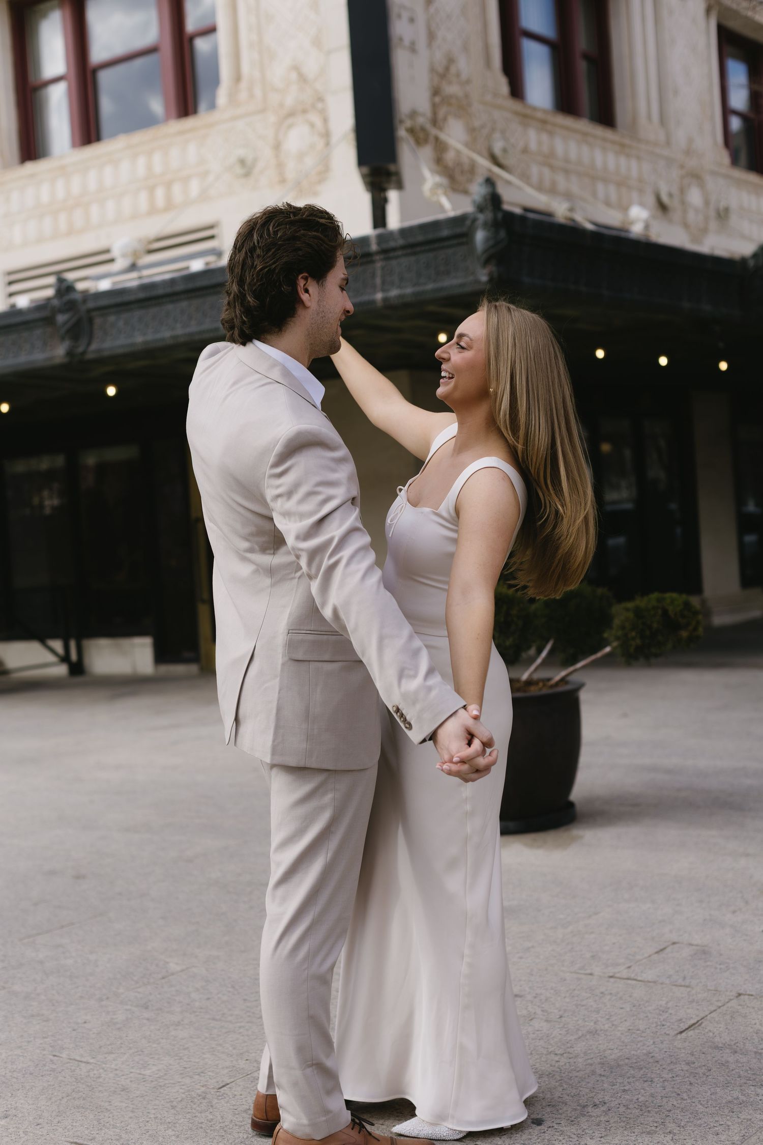 A couple in elegant white and beige attire share a romantic moment outside a historic building.