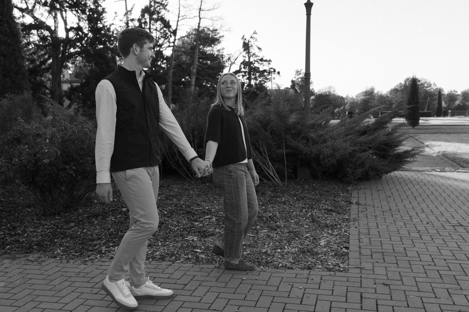 Black and white photo of couple walking together on brick pathway surrounded by landscaping.
