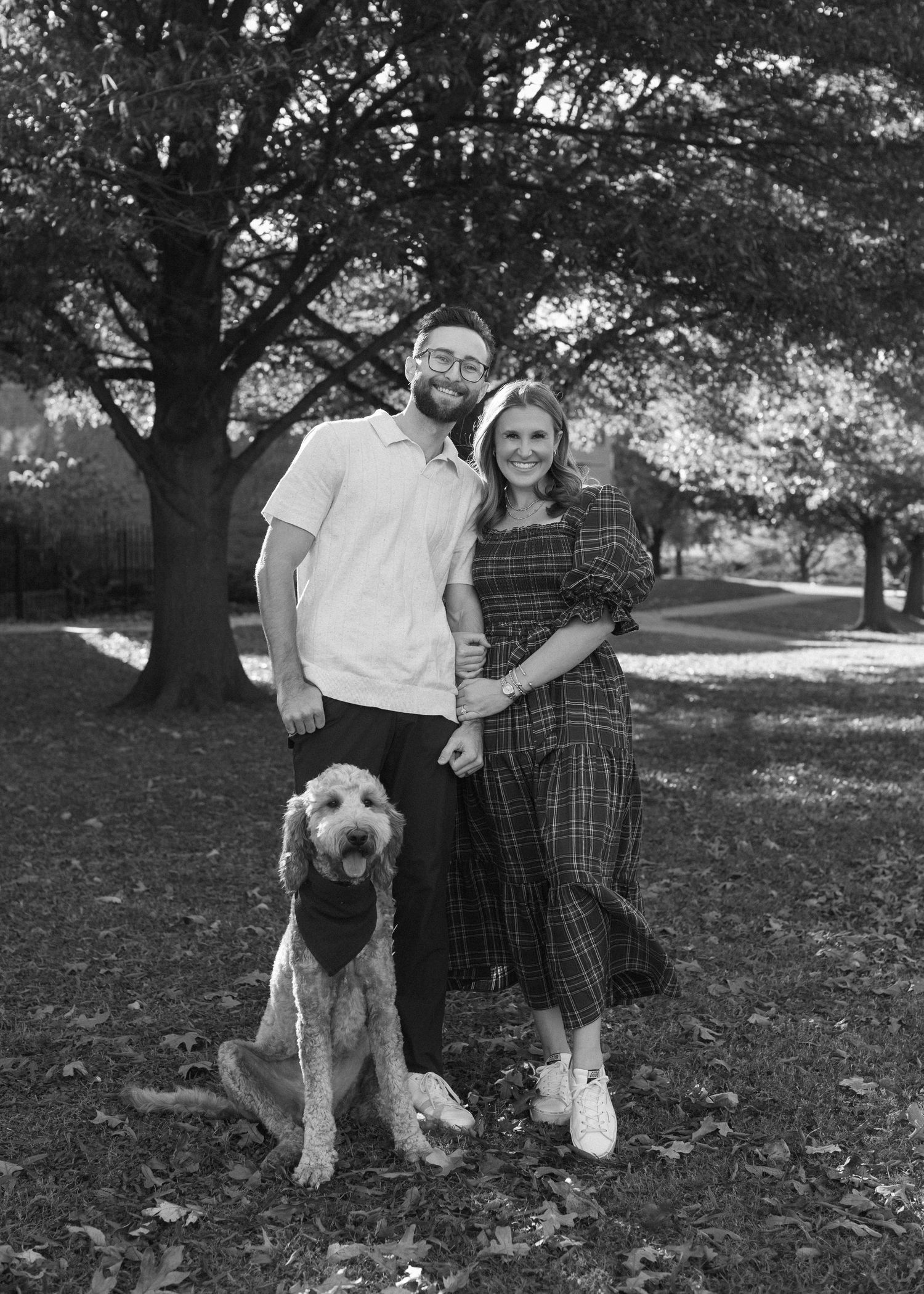 A couple poses with their goldendoodle dog in a park setting with large trees in black and white.