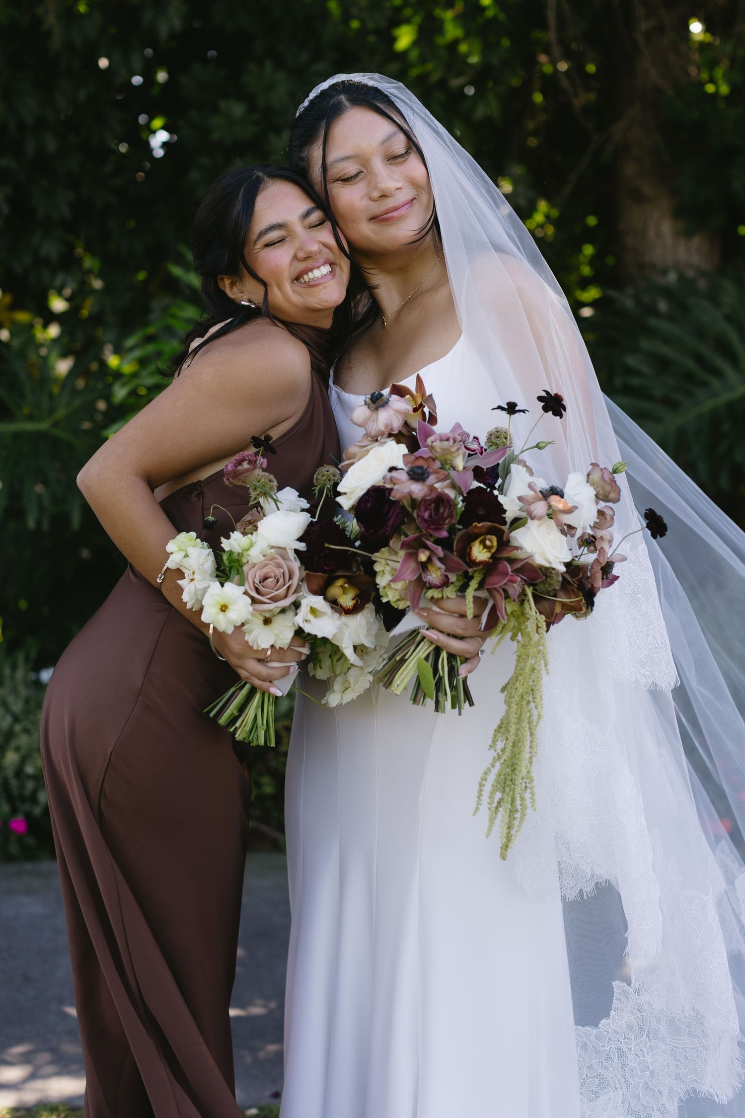 Wedding party members in brown dresses embrace the bride holding burgundy and white floral bouquets.