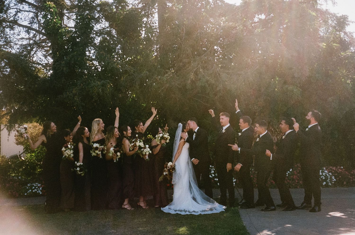 Wedding party celebrating outdoors with raised arms against a sunset backdrop.
