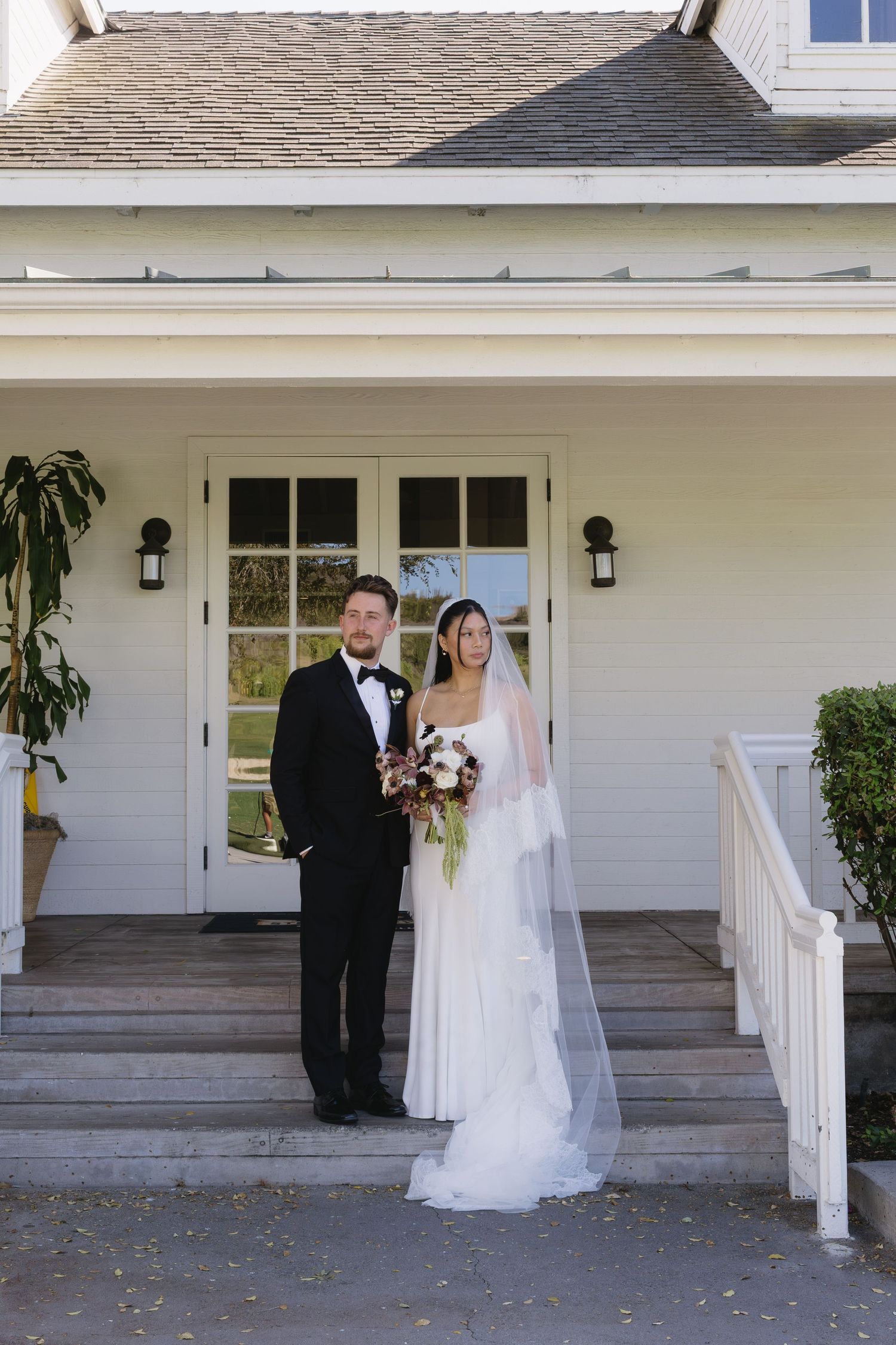 Wedding couple poses along white porch railing of classic farmhouse style building.