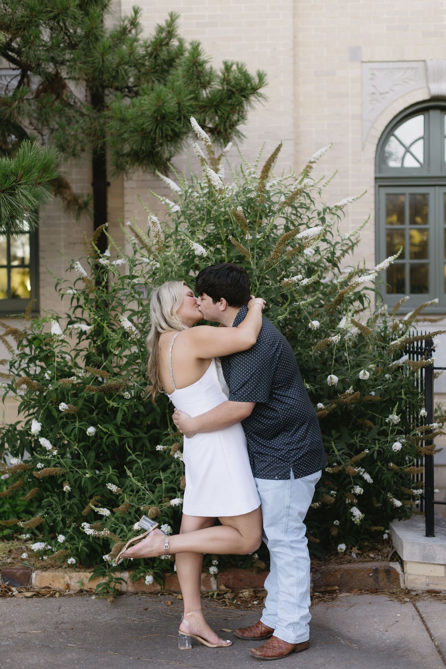 A romantic couple embraces in front of green foliage and building windows during a photo session.