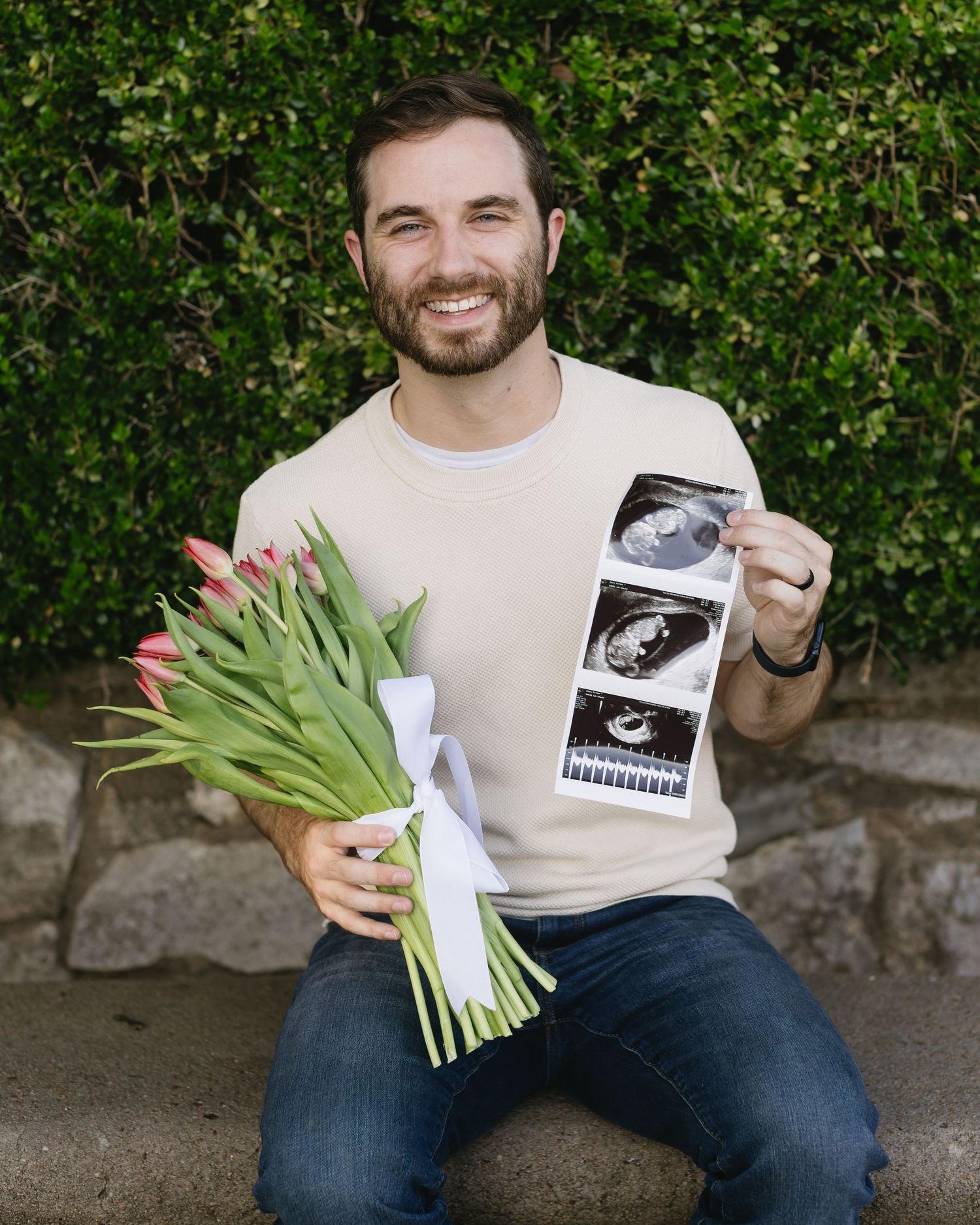 A person in a cream sweater displays a sonogram photo while holding tulips against a garden backdrop.