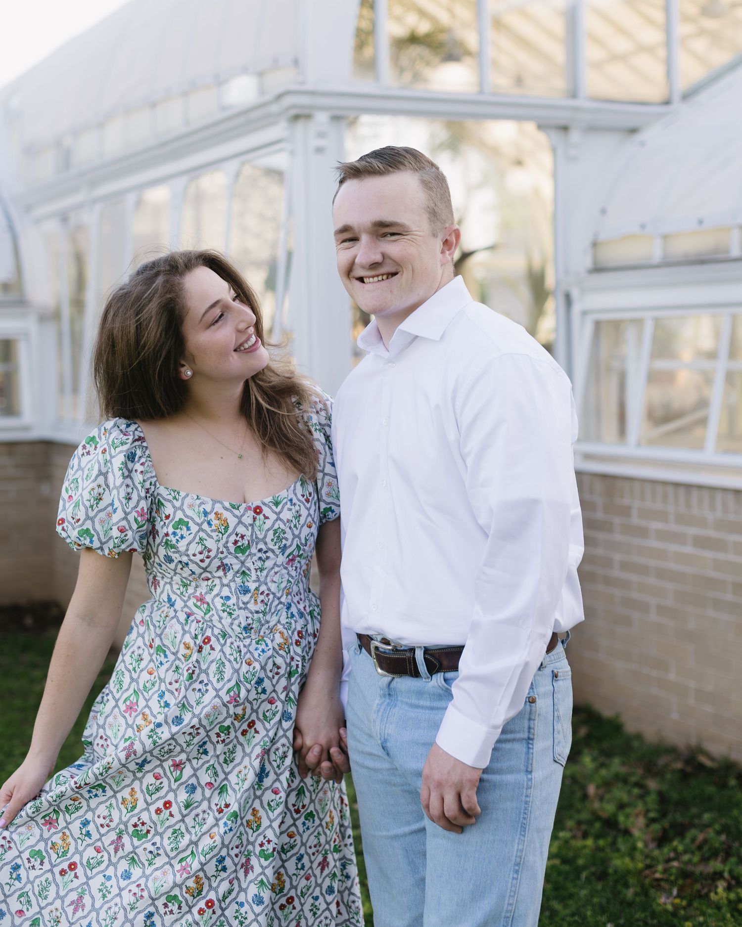 A couple poses together outdoors near a white greenhouse, wearing a floral dress and white shirt with jeans.