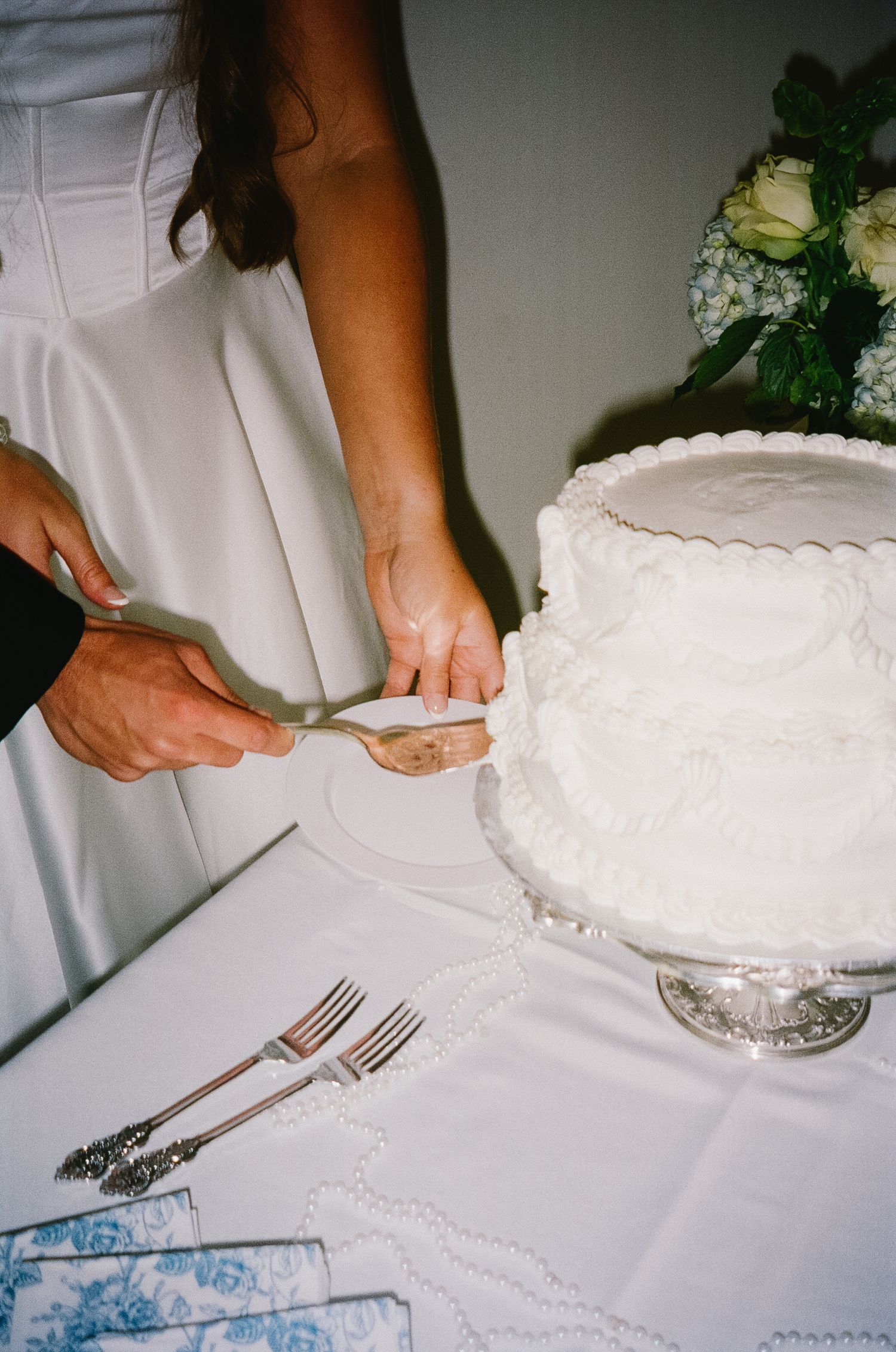 Someone in white clothing cuts into a traditional white wedding cake displayed on an elegant silver cake stand at a reception.