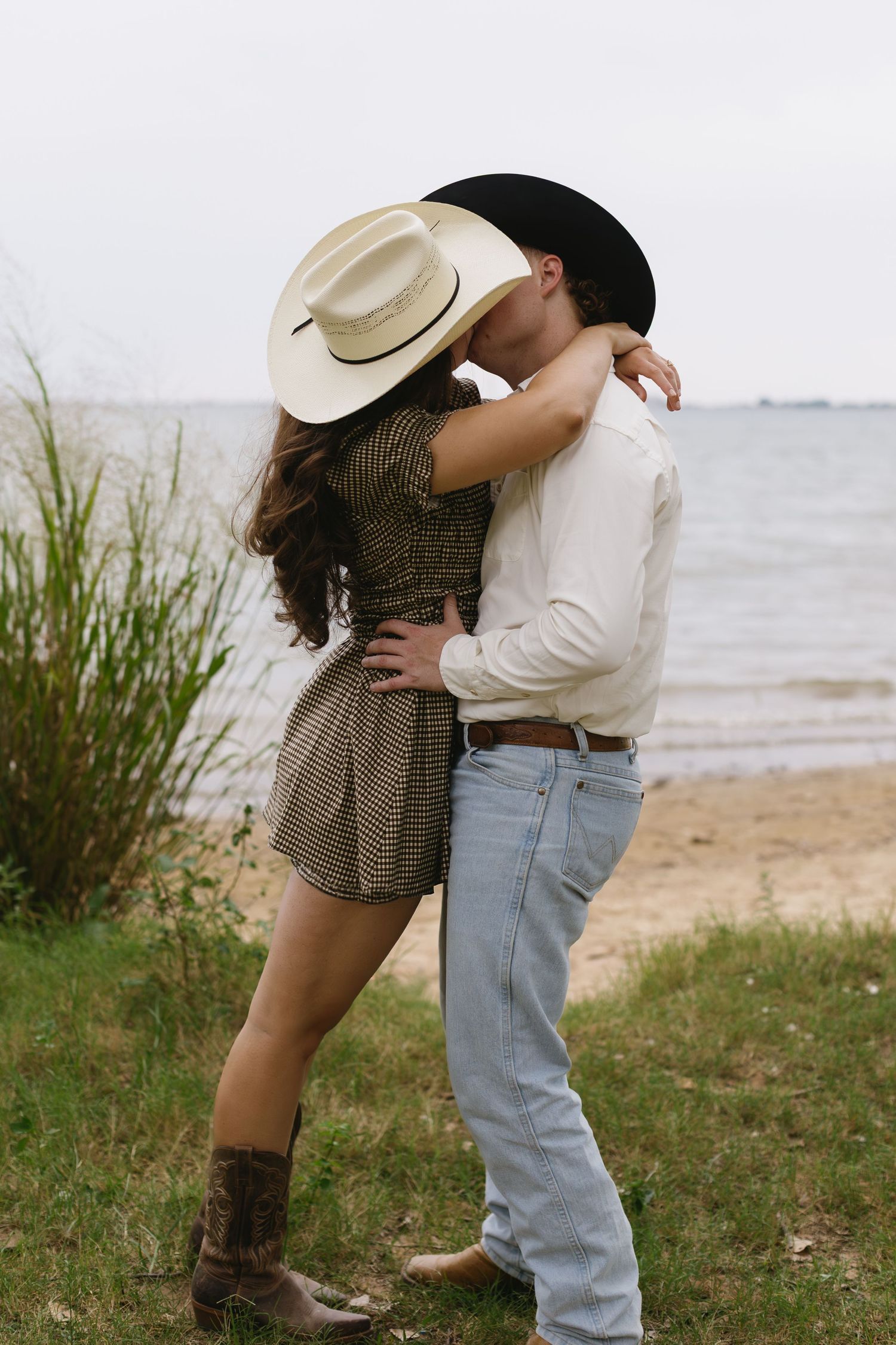 Romantic couple in western attire embraces near beach grass wearing cowboy hats.