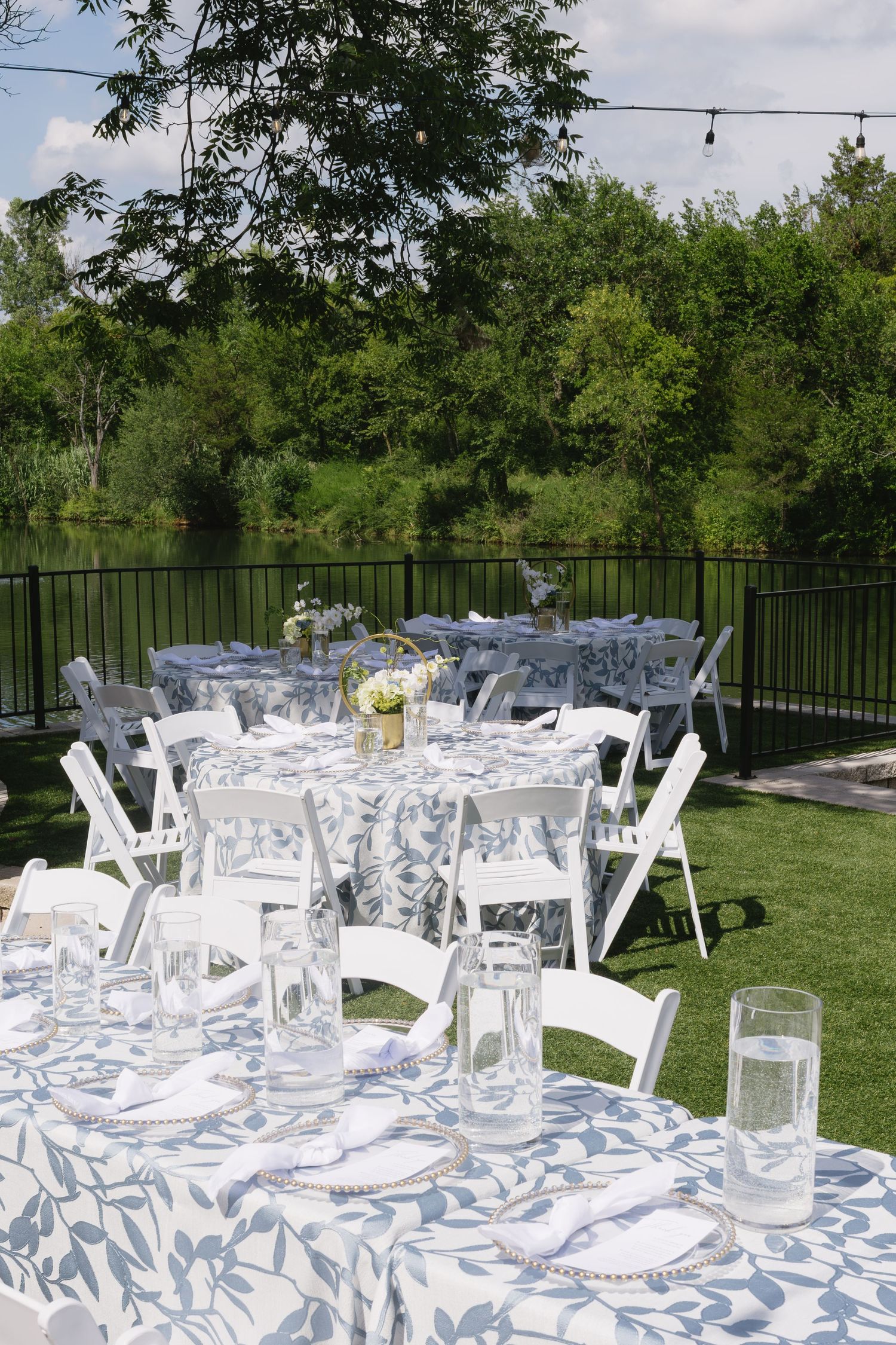Outdoor wedding reception setup with white folding chairs and tables arranged on a lawn surrounded by lush greenery.