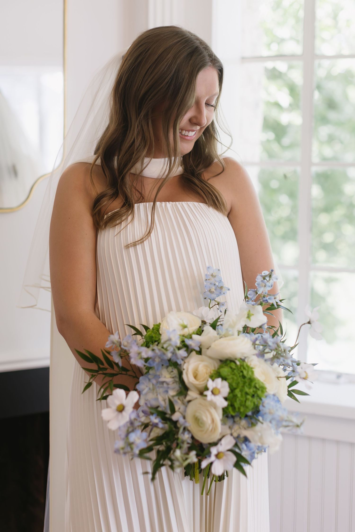 A person in a white pleated dress holds a beautiful bouquet of white roses and blue delphinium flowers by a window.