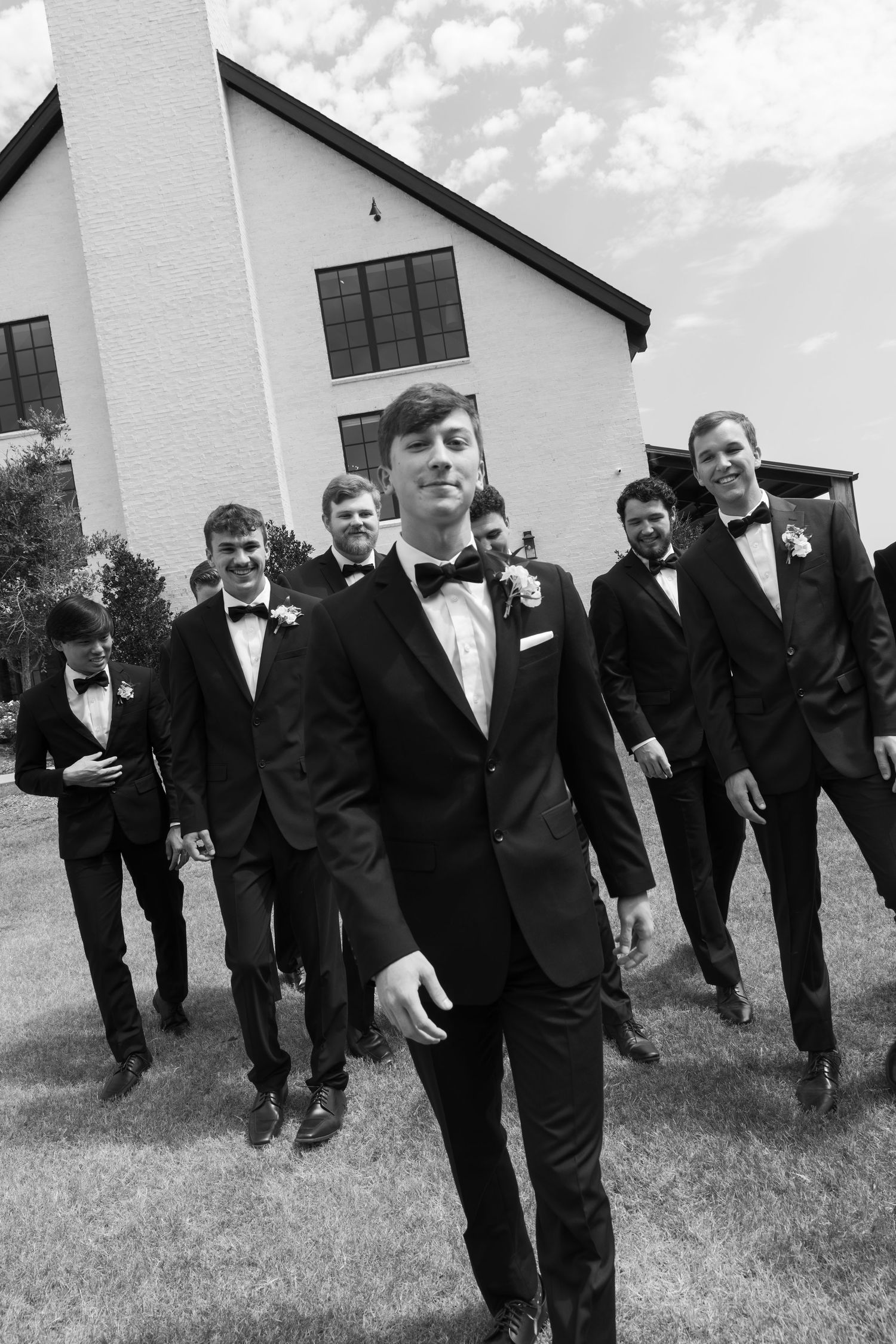 Groomsmen in black tuxedos posing playfully outside a white church building.