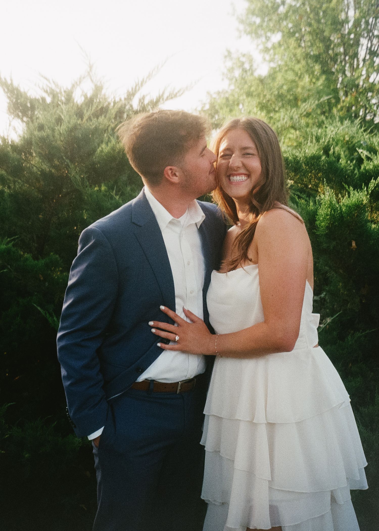 A couple shares a tender moment in a garden setting wearing wedding attire during a sunny outdoor photo session.