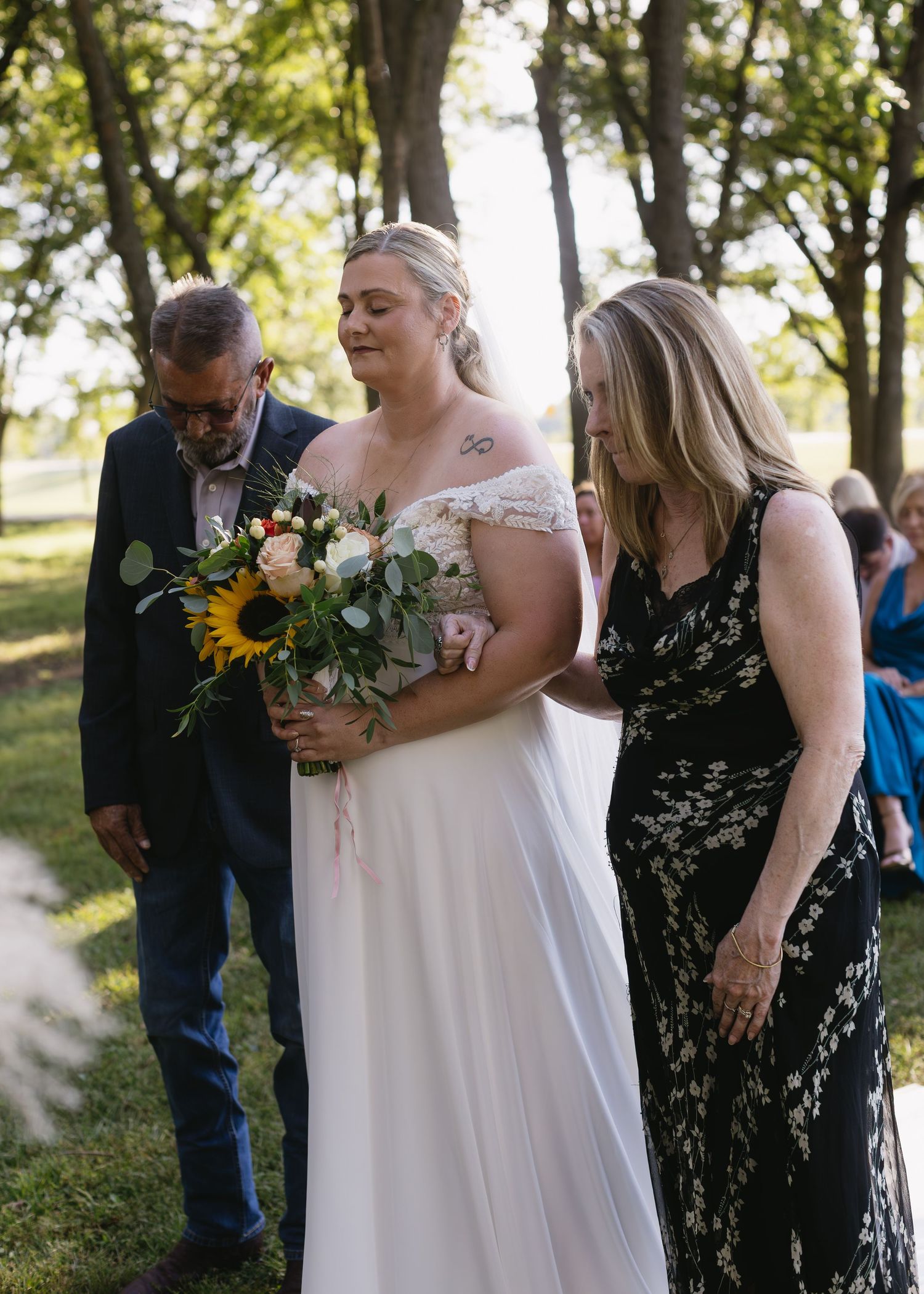 A wedding ceremony takes place outdoors with attendees gathered under trees.