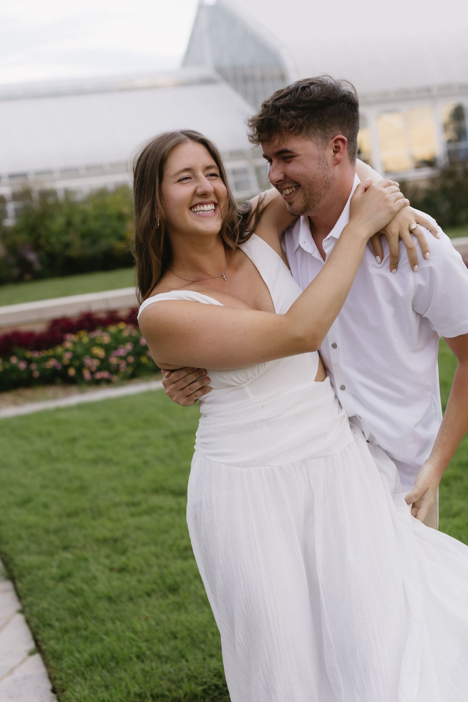 A playful couple laughing and embracing on a manicured lawn with flowers in the background.