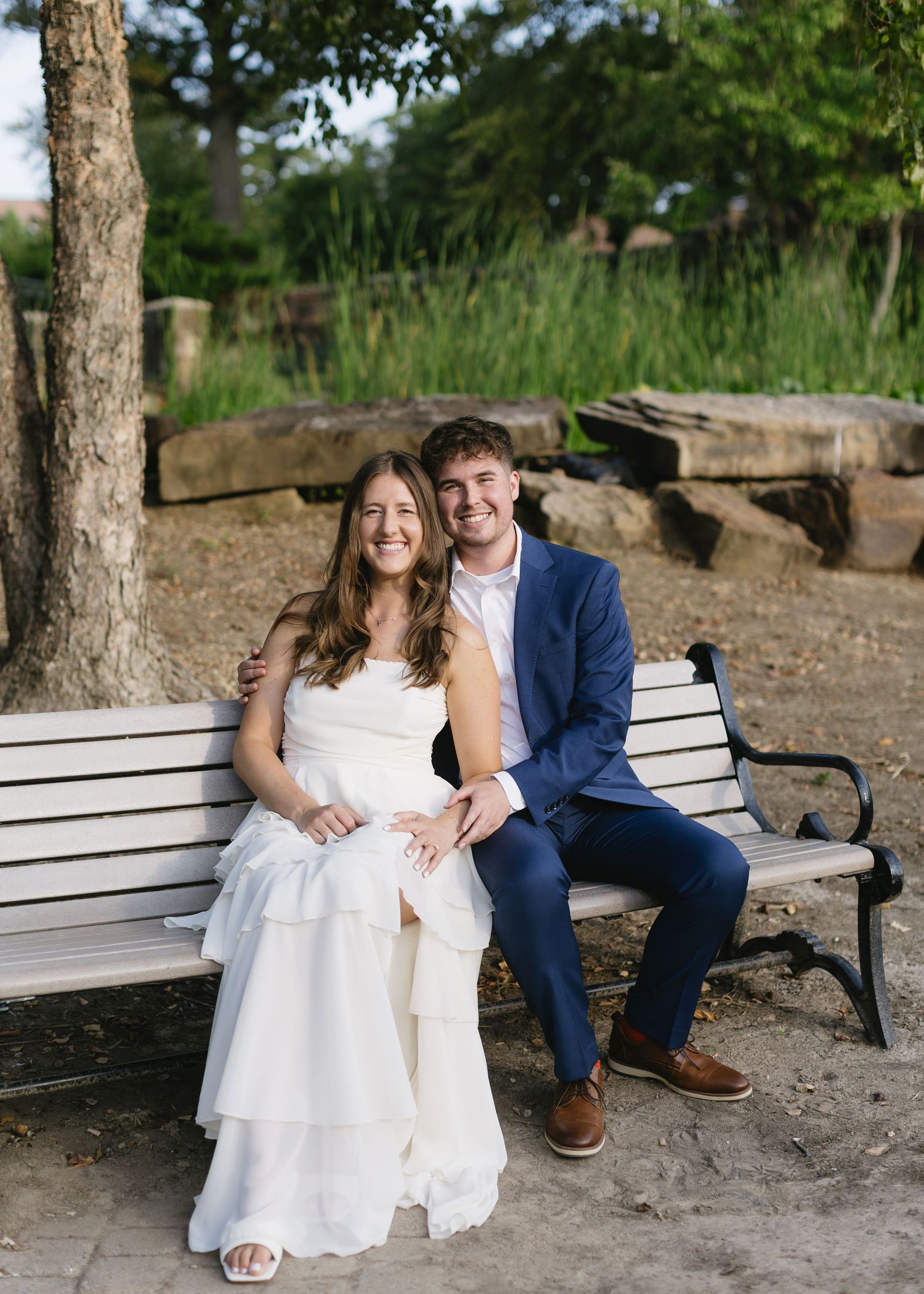 A couple sits together on a park bench surrounded by trees and natural lighting.