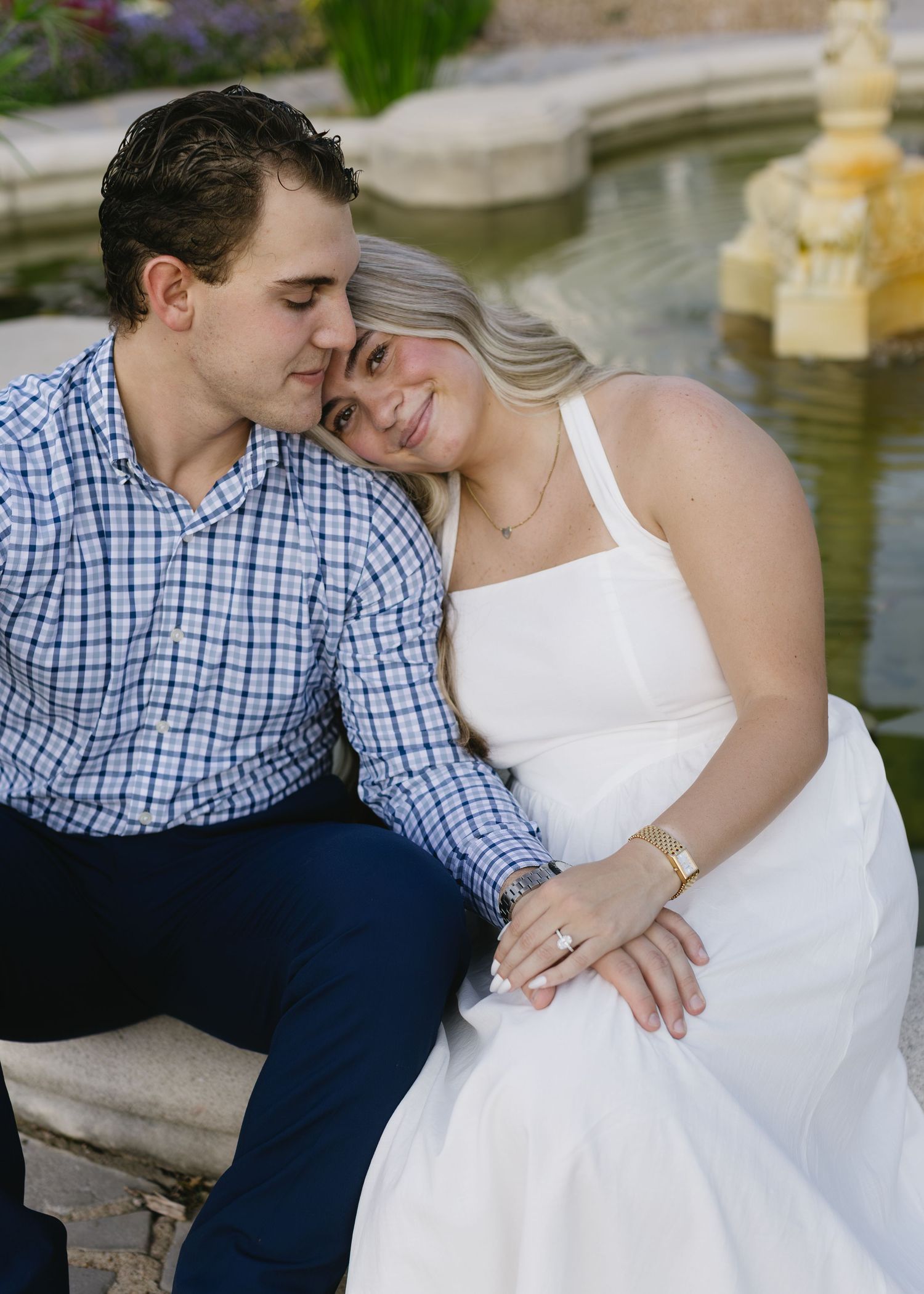 A couple in blue plaid shirt and white dress embraces near a decorative fountain.