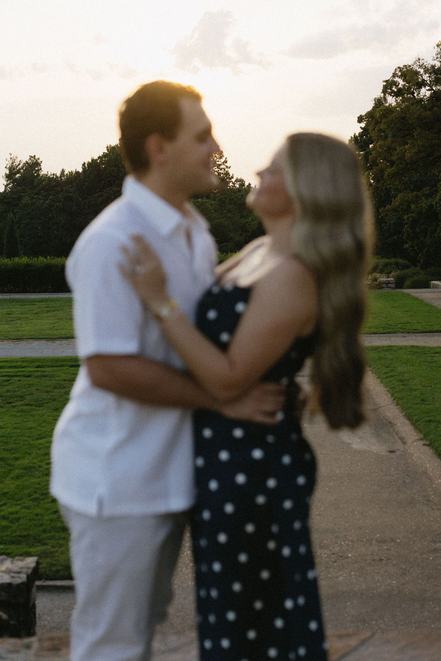 Couple dancing and embracing outdoors on a paved path with green trees and grass in the background.