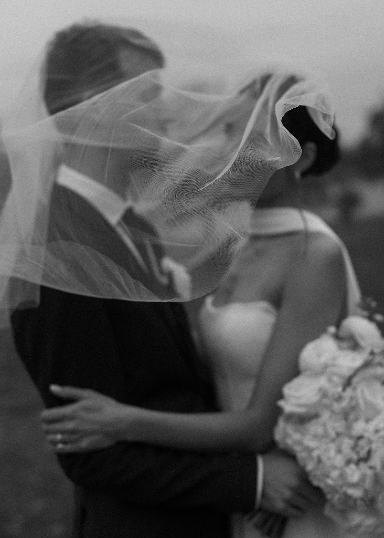 Romantic black and white photo of newlyweds embracing under a flowing veil.