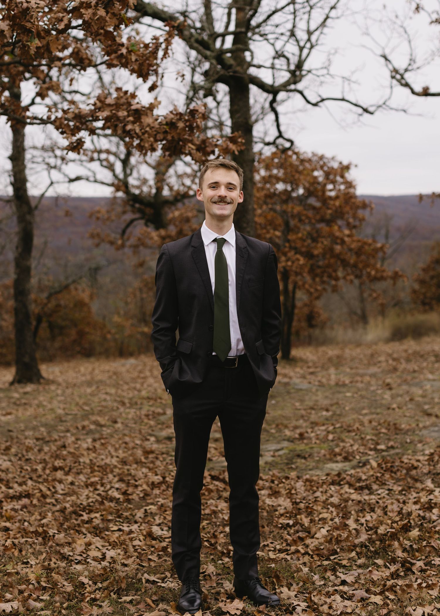 A black suited figure poses in a woodland setting with fallen autumn leaves.