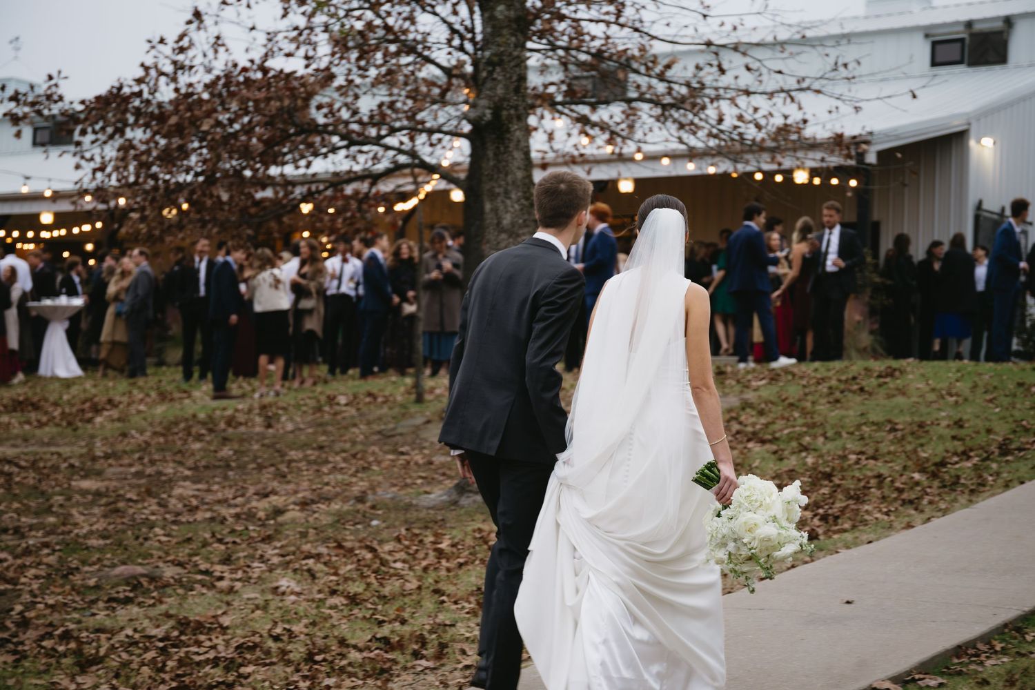 Newlyweds walking along path towards reception venue with string lights and guests in background.