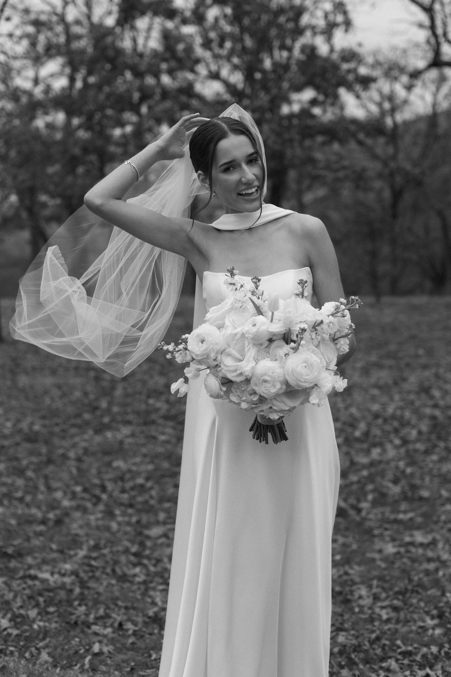 Elegant bride in a flowing white dress holds a lush bouquet of white flowers in an outdoor setting.