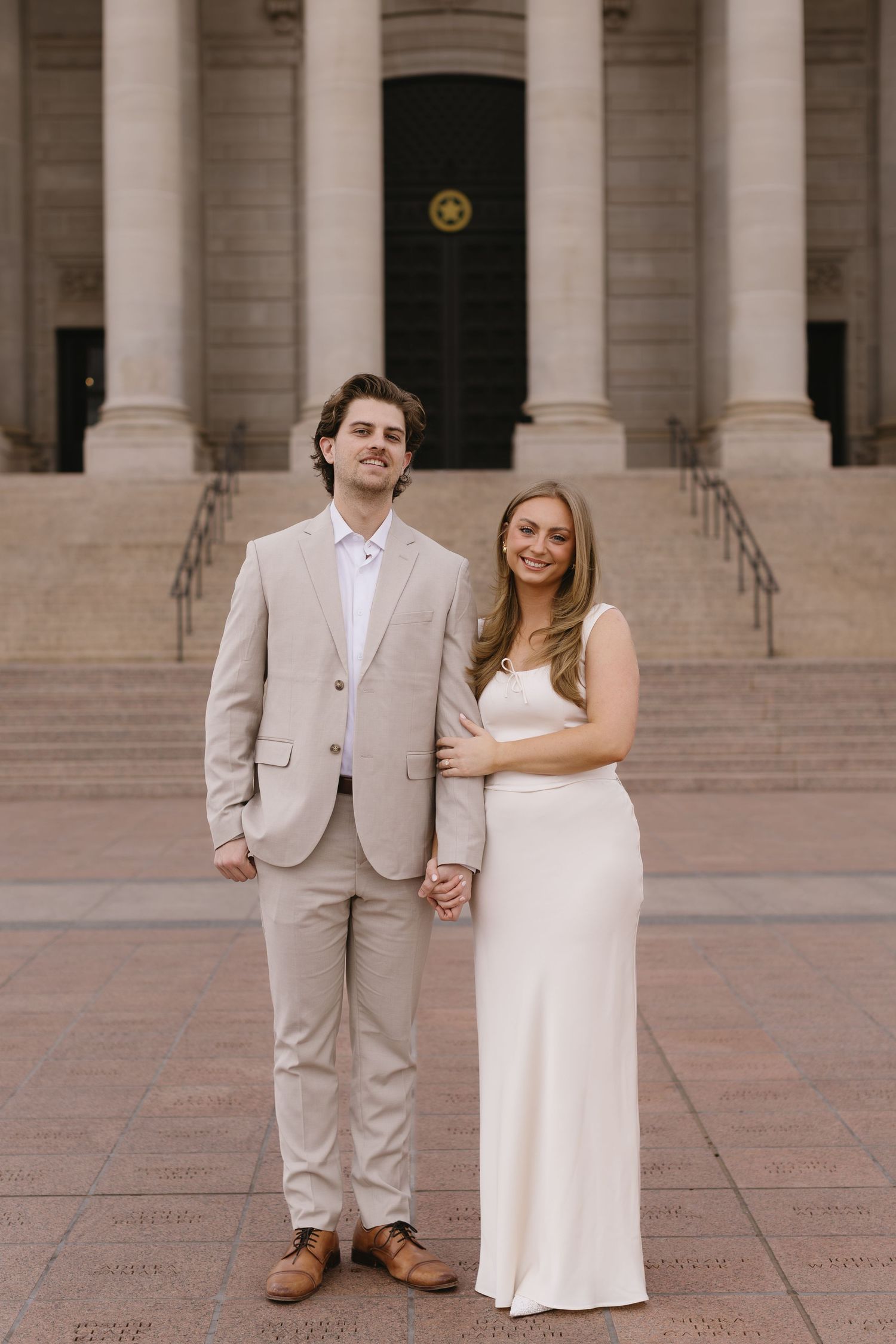 A couple in formal attire poses on stone steps of a grand building with columns.