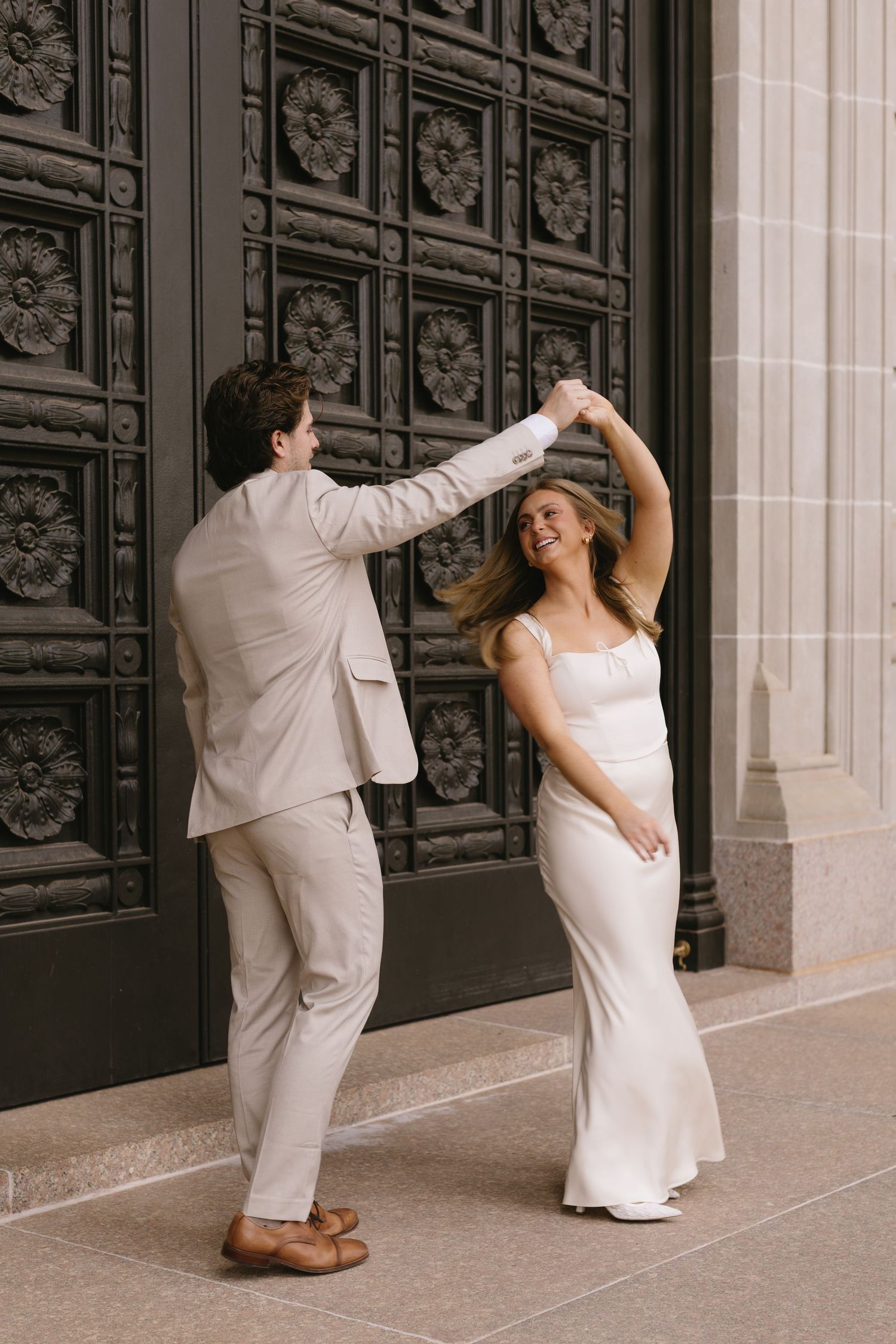 A playful couple in formal wear dances together near ornate black doors.