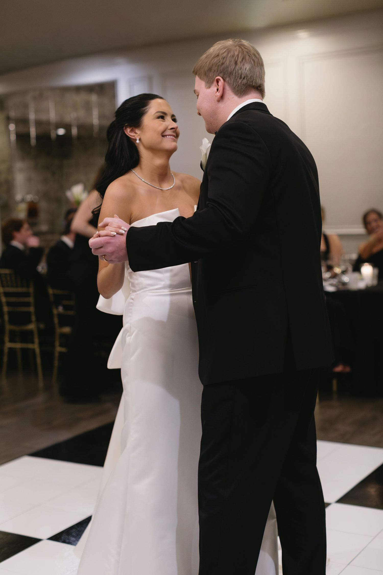 Elegant couple shares their first dance in a white dress and black tuxedo at indoor reception.